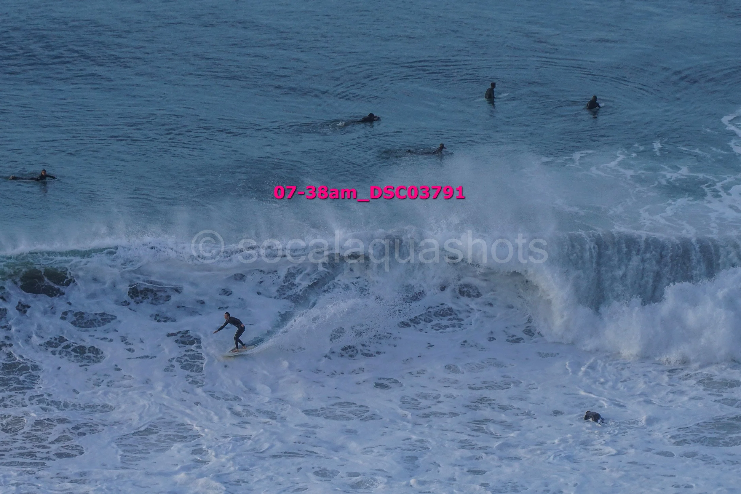 A person surfing on a wave in the ocean with several other people swimming and floating in the water.