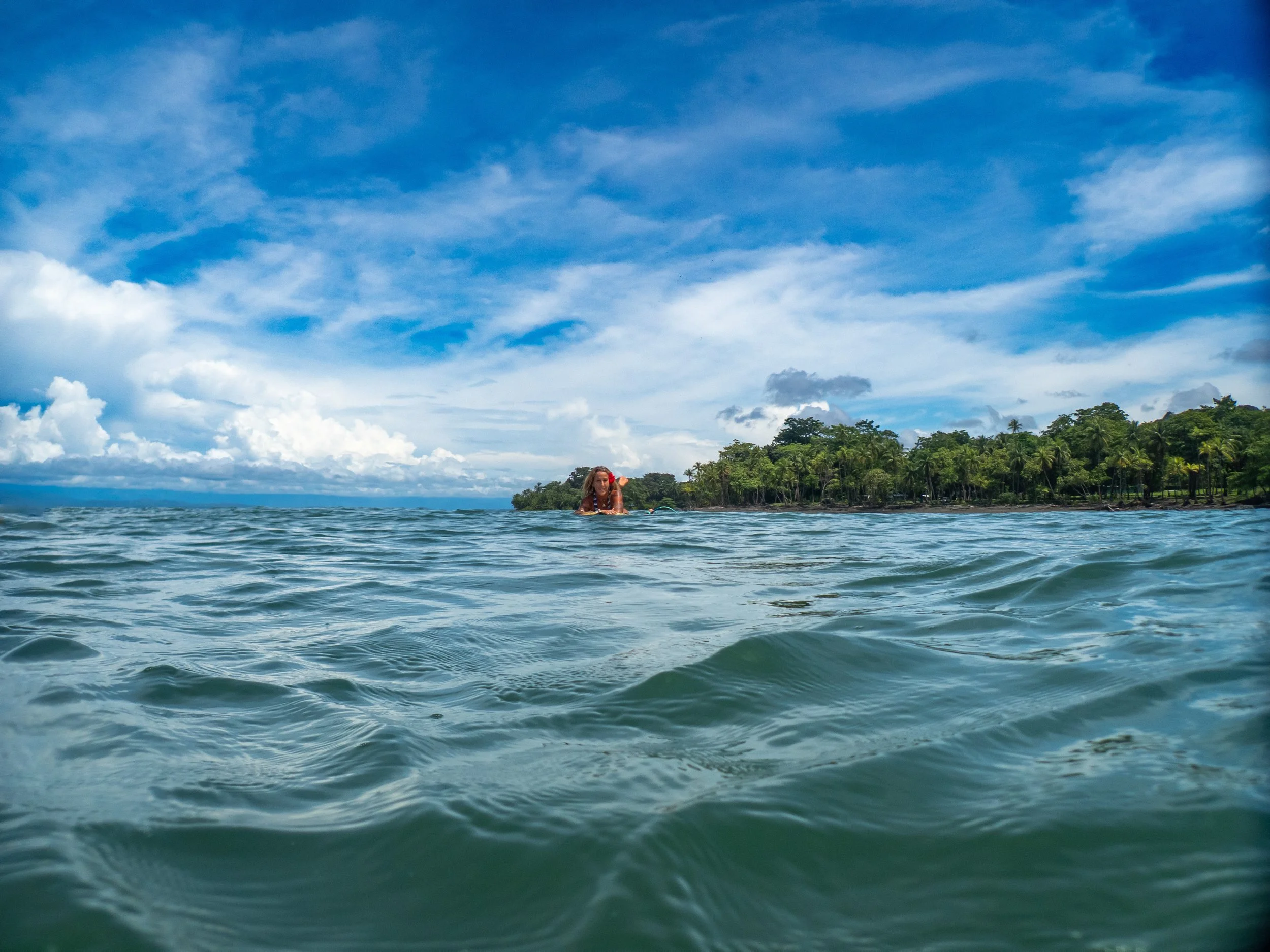 Person swimming in the ocean near a tropical island with lush green trees and a partly cloudy sky.