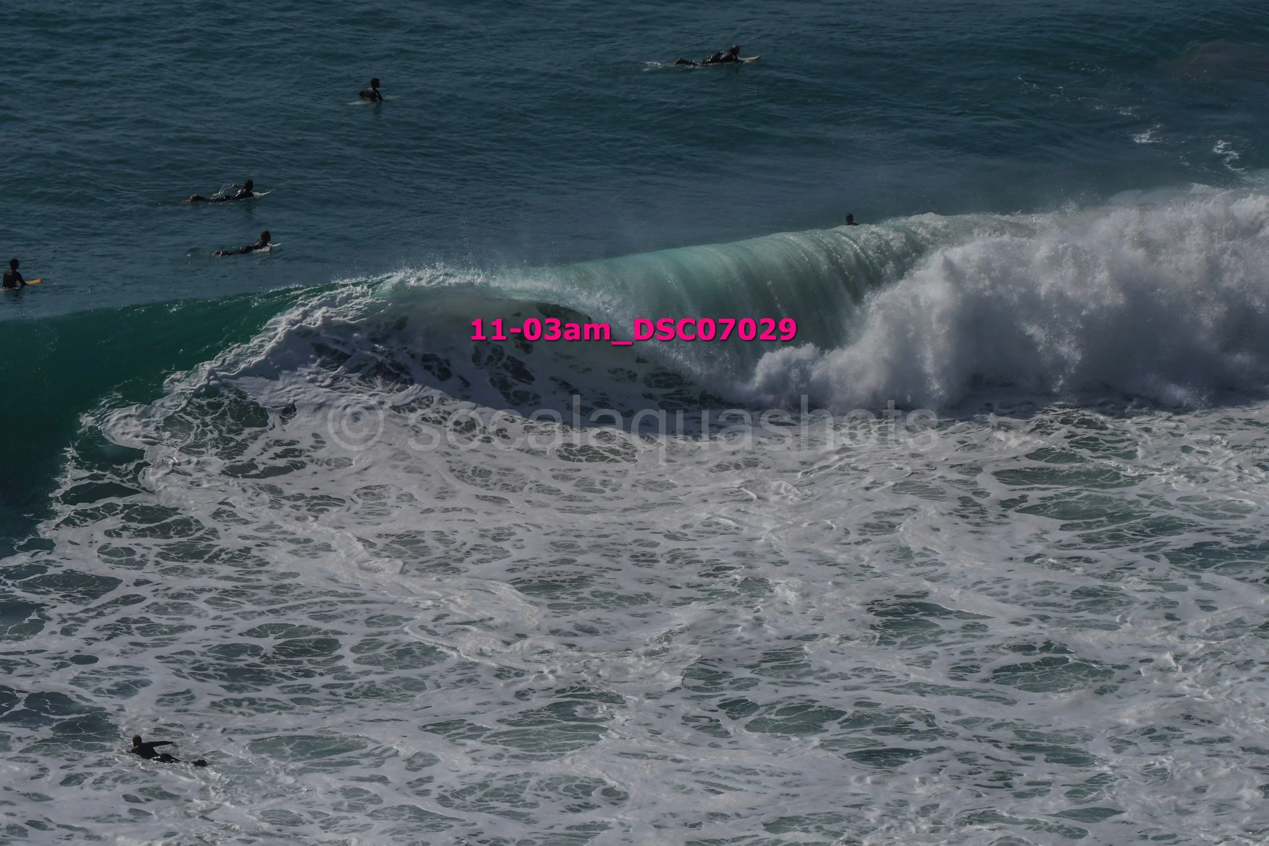 Multiple surfers in the ocean, some riding a large wave, with others waiting in the water, under a clear sky.