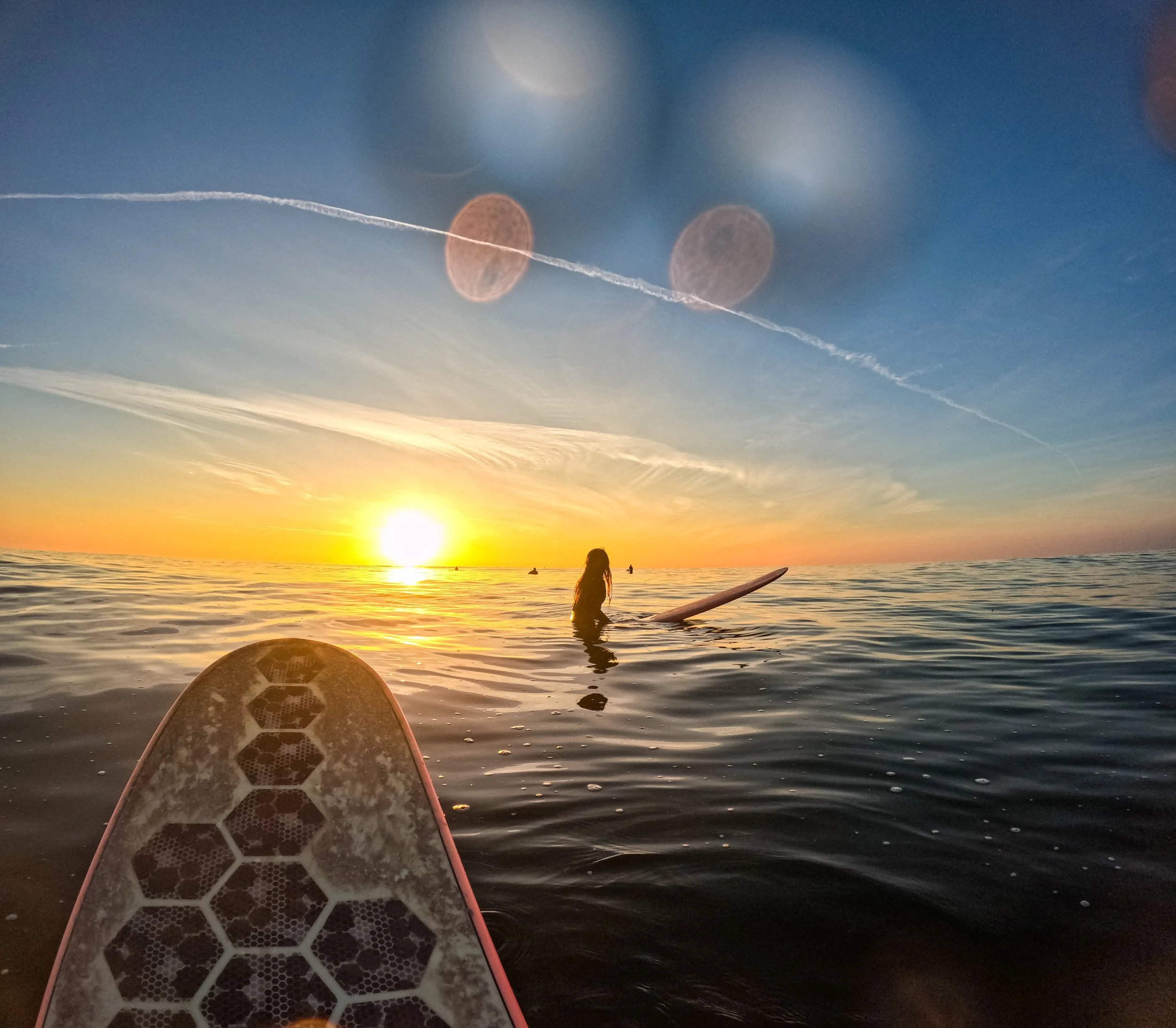 View from a paddleboard in the ocean during sunset, with a person standing on a surfboard in the water, and the setting sun on the horizon. The sky has clouds and contrails, with lens flare present.