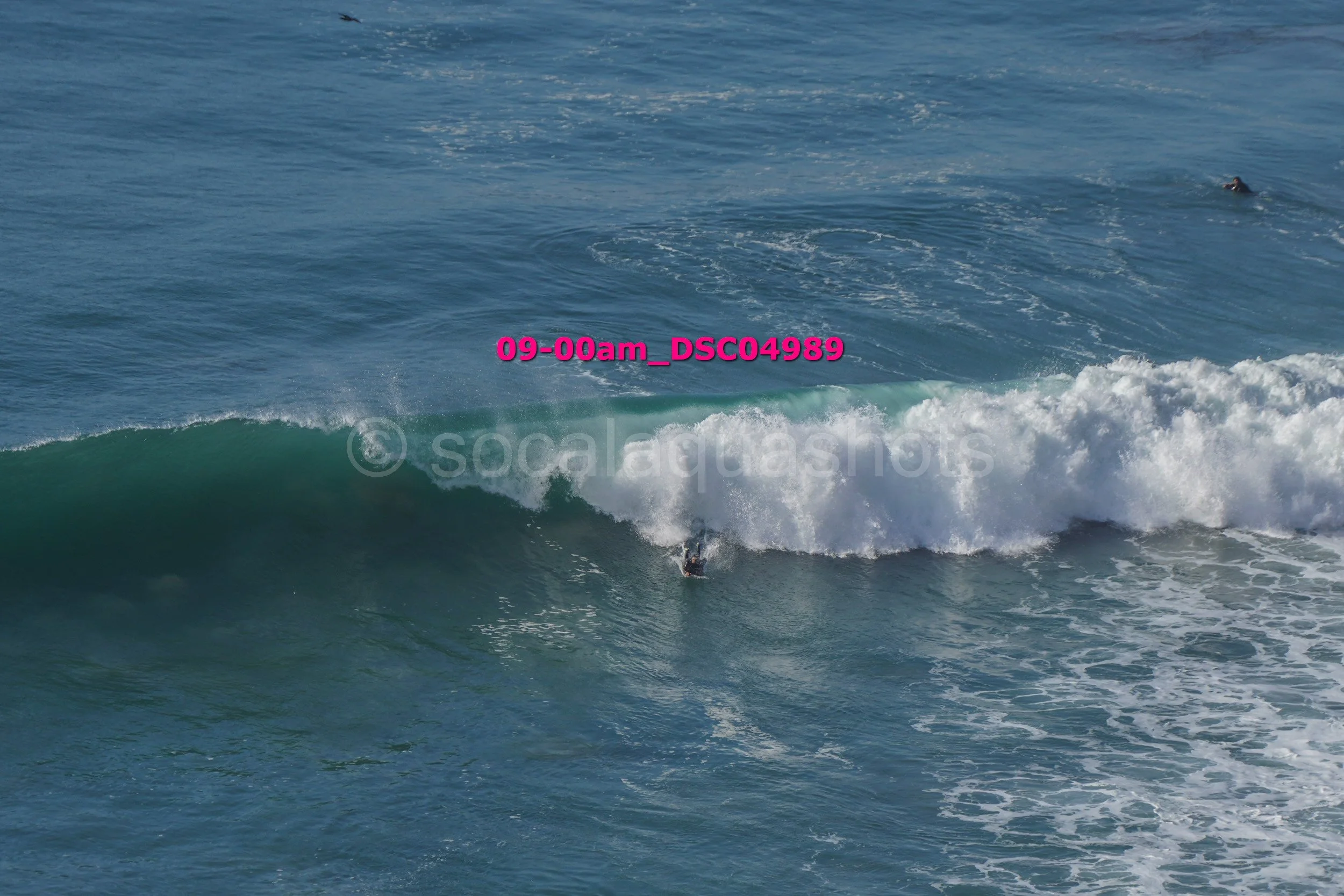 A person surfing on a large ocean wave, with another swimmer visible in the distance.