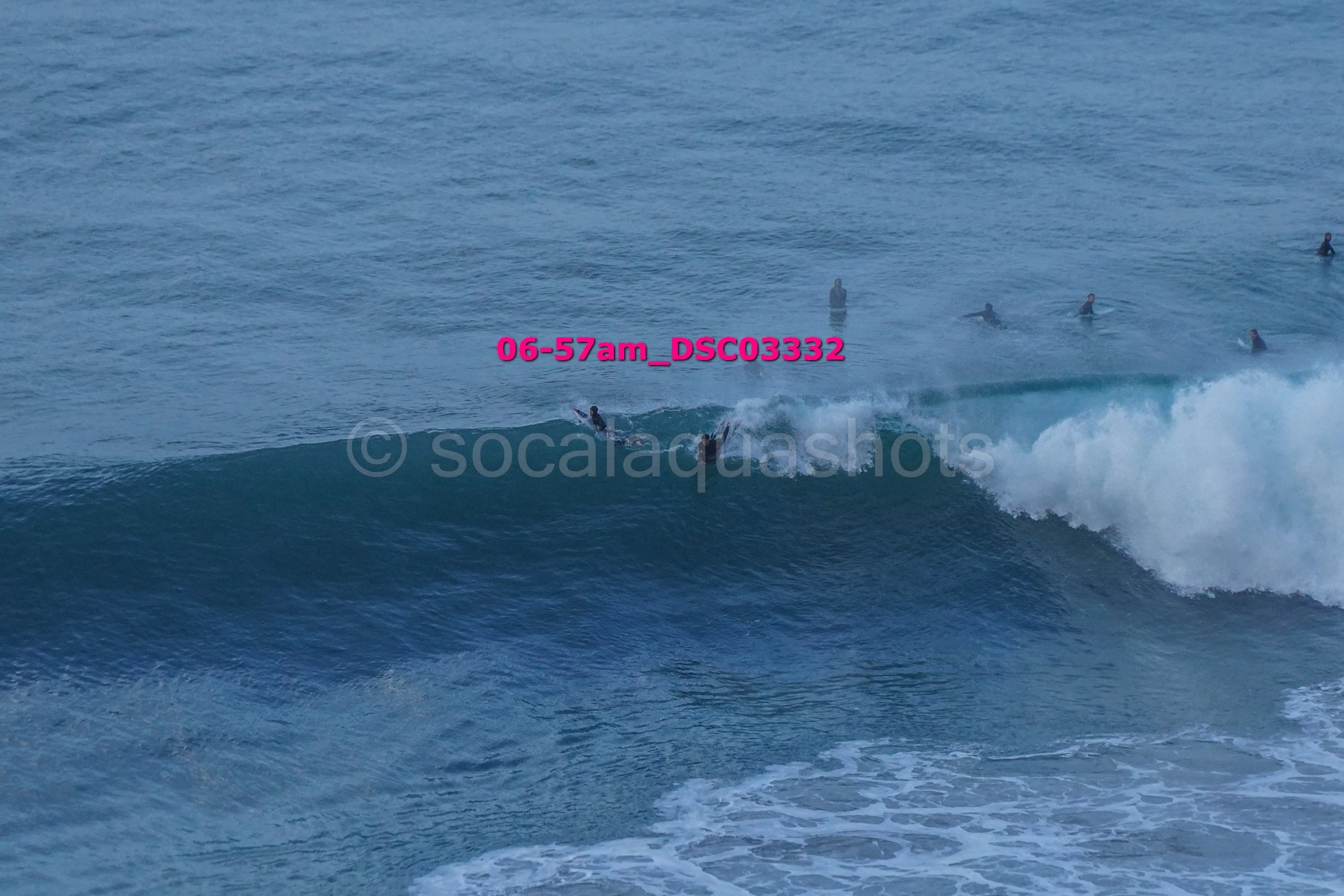 A group of surfers riding a wave in the ocean, with some surfers visible in the water in the background.