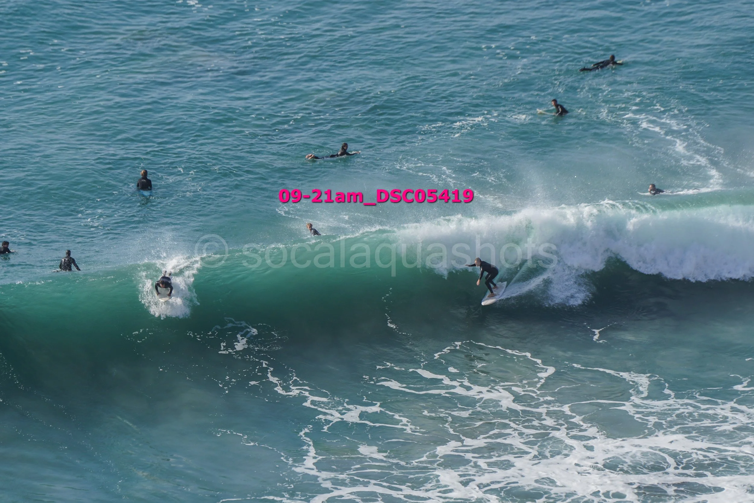 Group of surfers riding and waiting for waves in the ocean, with some on surfboards on a large wave and others floating in the water.