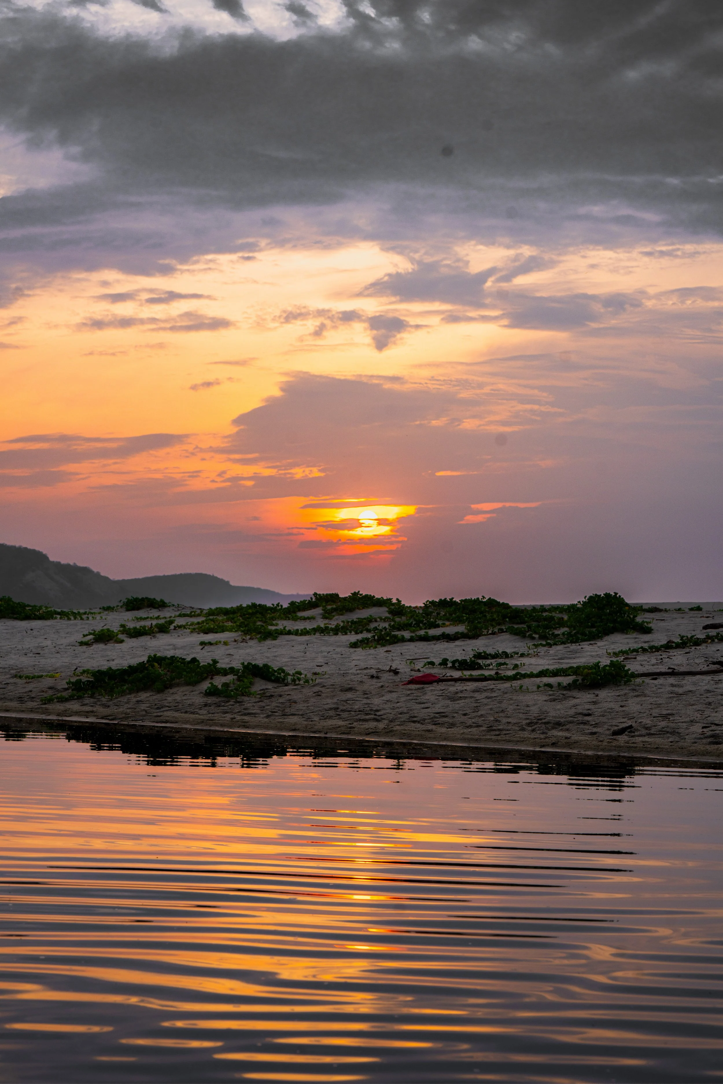 A sunset over a body of water with reflections, distant hills, and a cloudy sky.