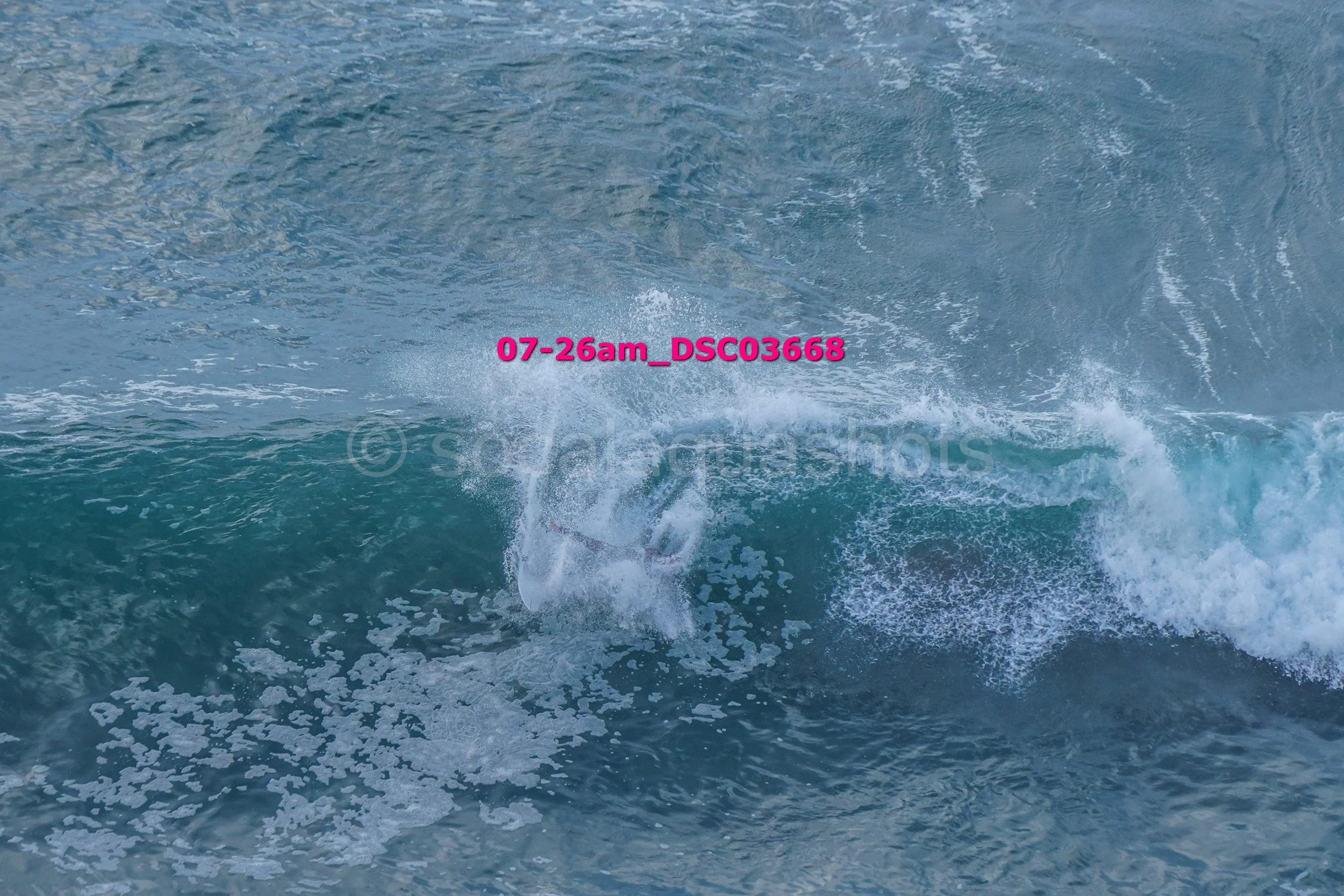 A large wave crashing in the ocean with white foam, with some pink text overlay reading '07-26am_DSC3668'.