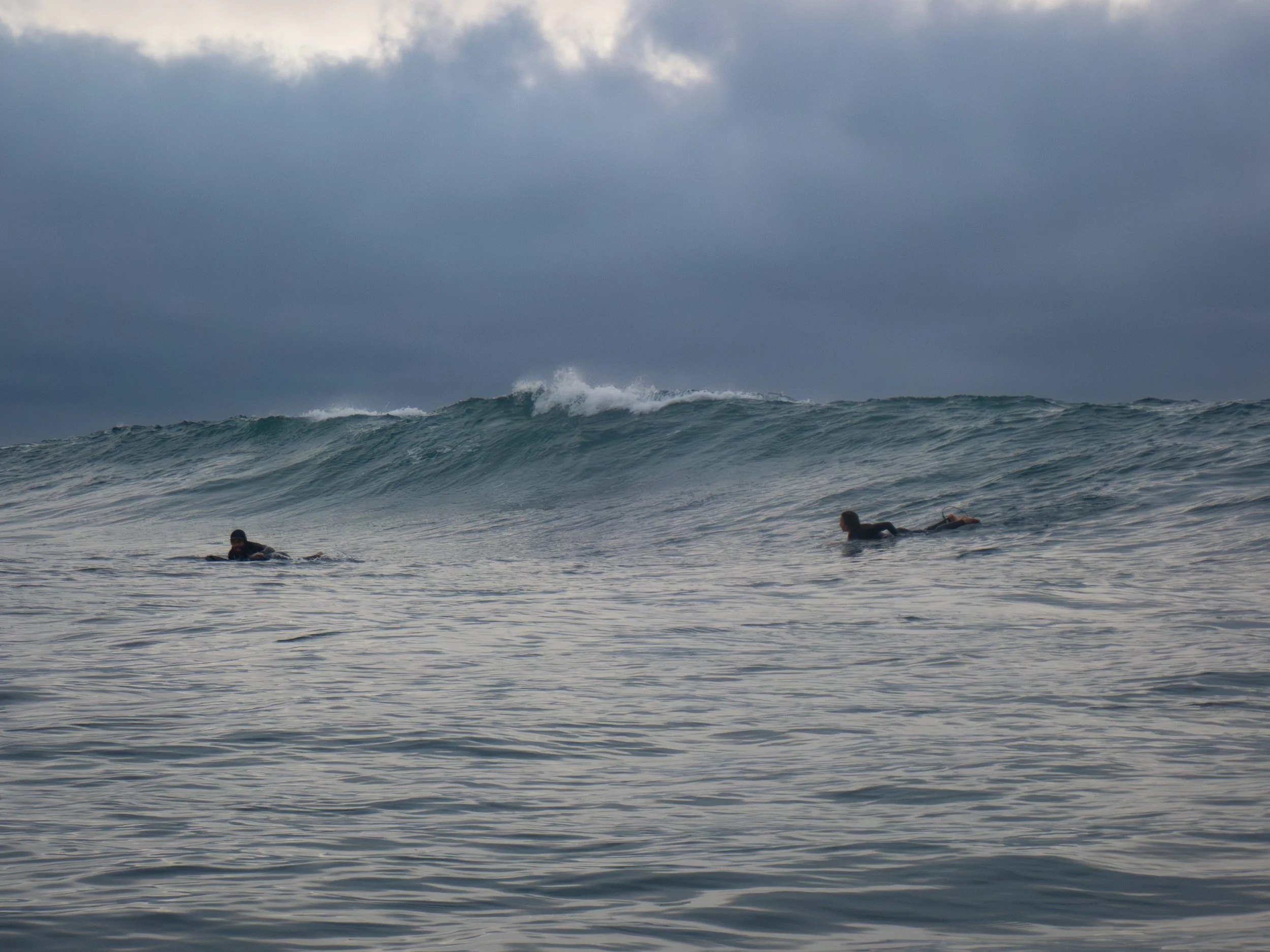 Two surfers in wetsuits paddling on surfboards waiting for waves in an ocean with large rolling waves under a cloudy sky.