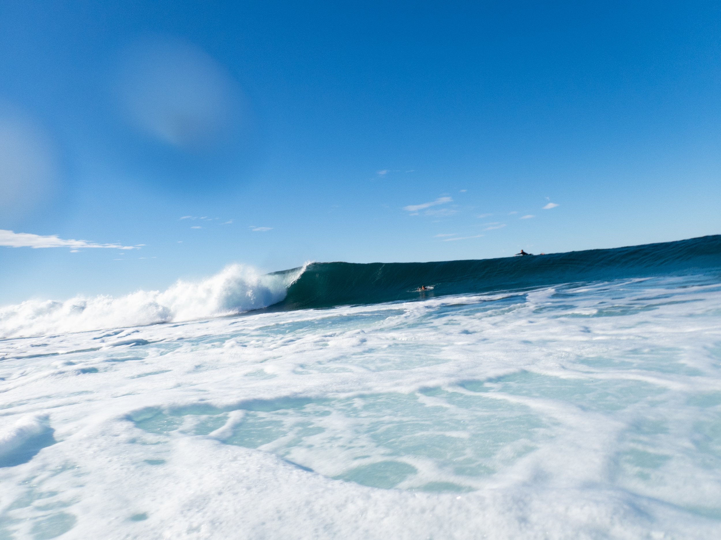 Ocean waves with clear blue sky, a person swimming in the water, and a boat in the distance.