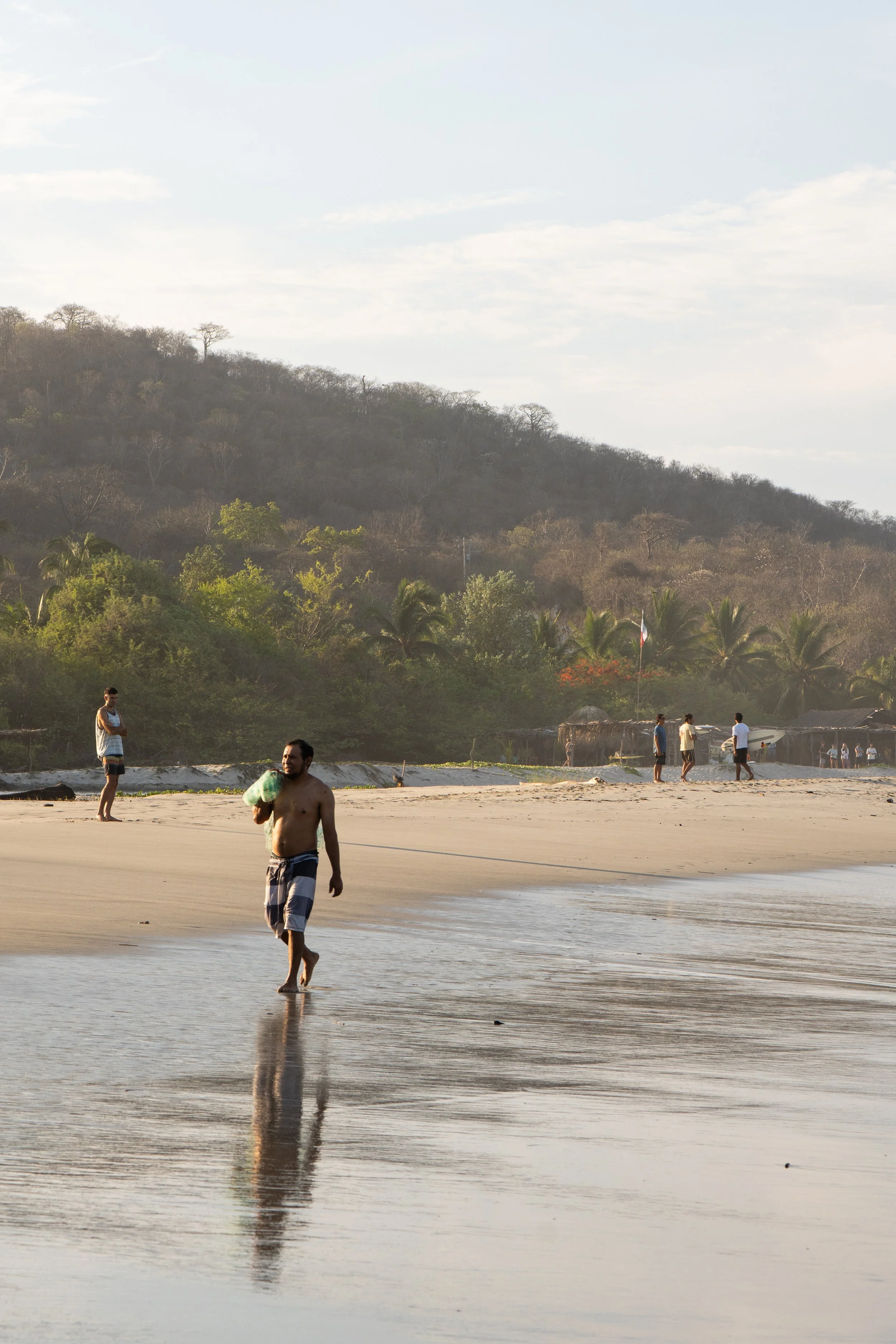 People walking on a sandy beach with a hillside covered with trees in the background. The sky is partly cloudy, and the water reflects the scene.