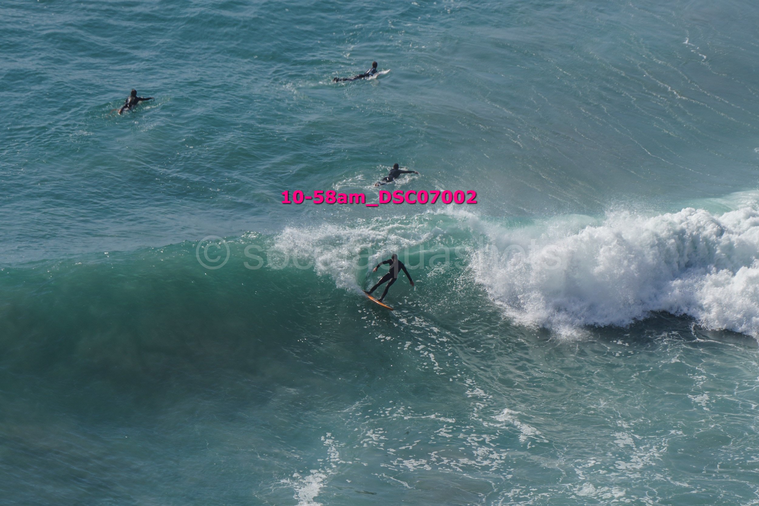 Surfing scene with one surfer riding a wave and three other surfers paddling in the water nearby.