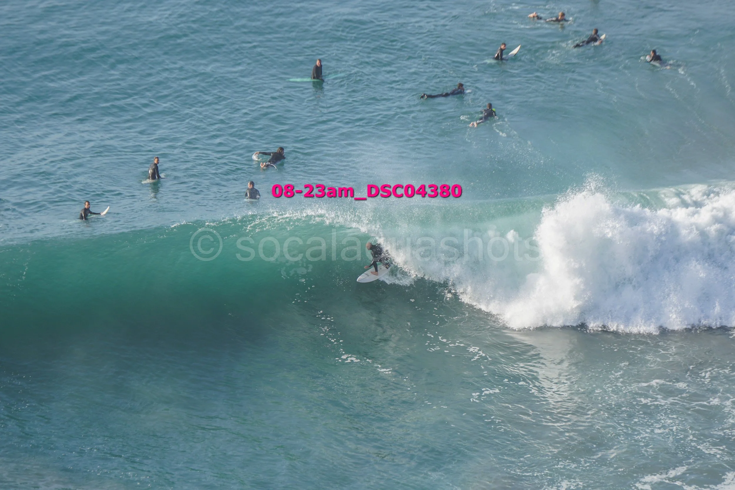 Surfer riding a wave with multiple surfers in the water in the background, some lying and some paddling on surfboards.