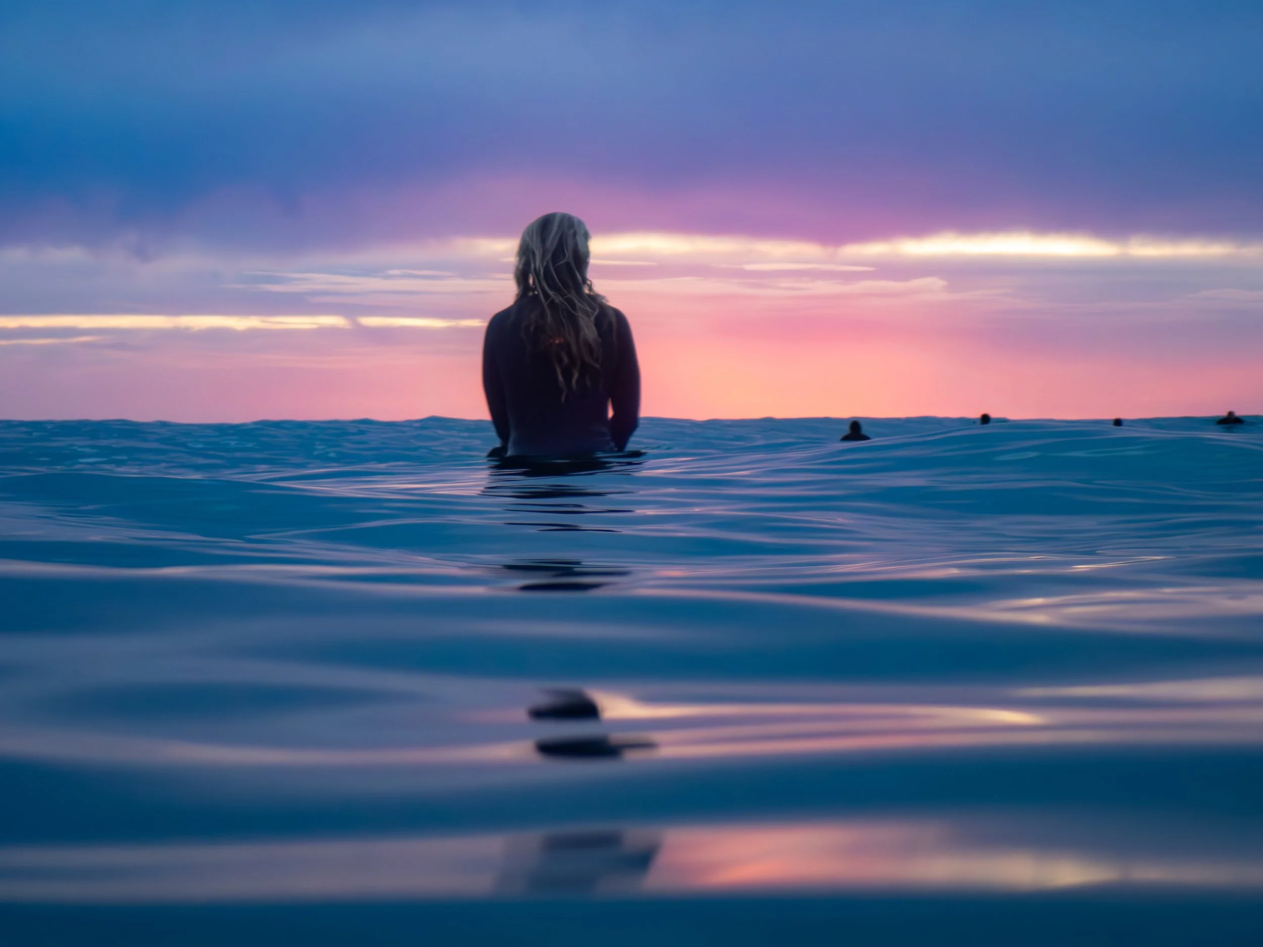 A woman with long hair sitting in the ocean during sunset, facing away towards the horizon with clouds in the sky.