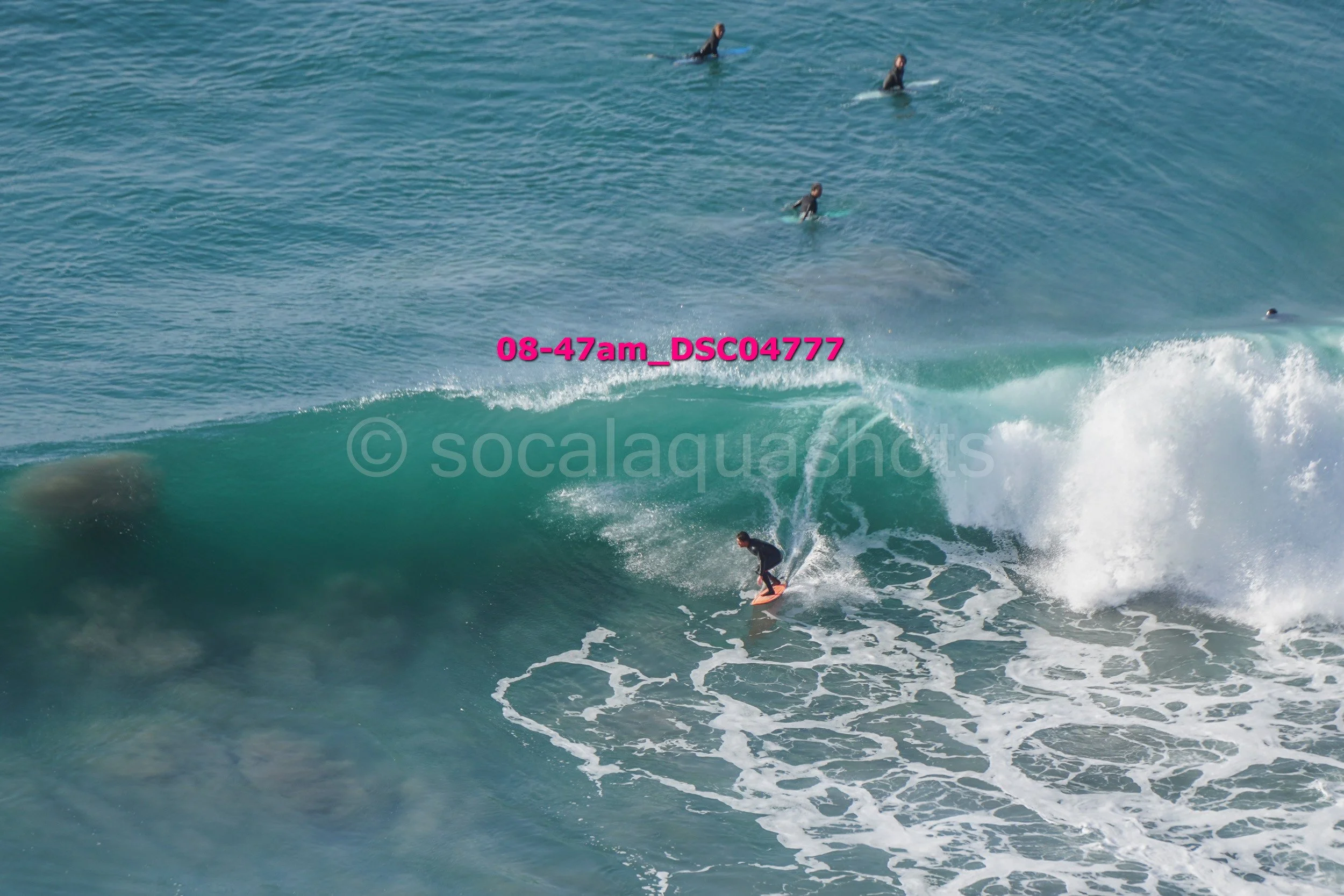A person surfing on a large wave while other surfers wait in the water nearby.