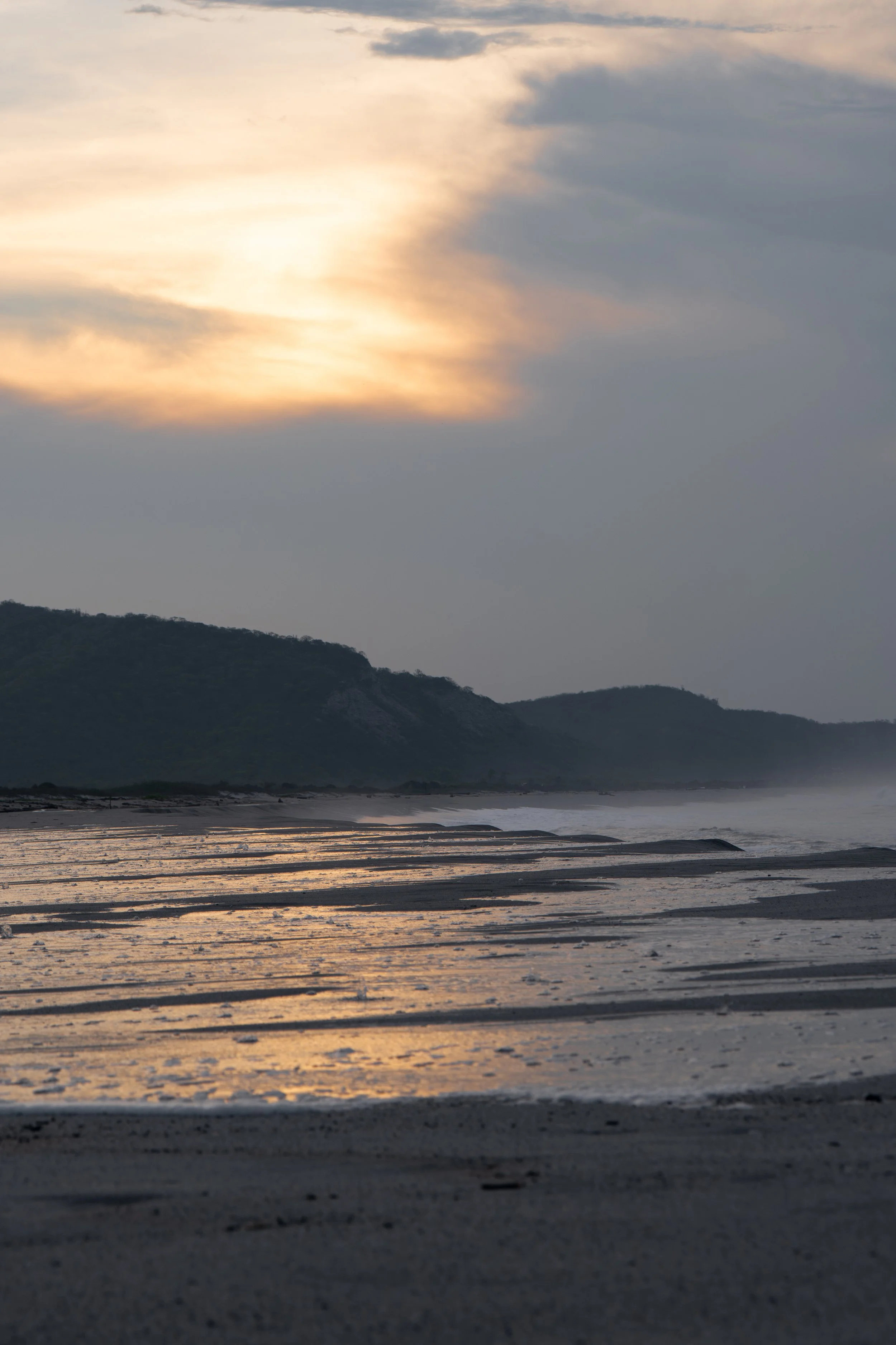 Sunset over the ocean with scattered clouds, distant mountains, and wet sandy beach foreground.