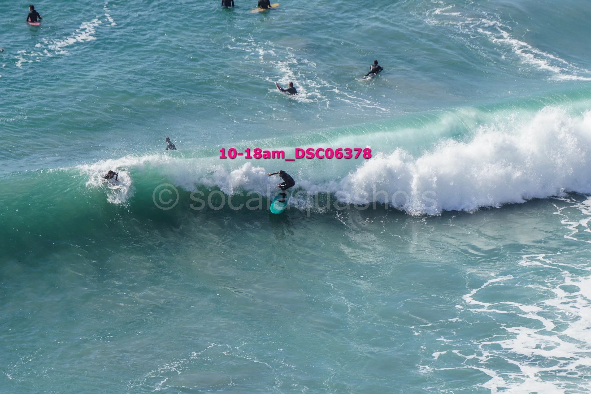 A person surfing on a turquoise wave at a beach while several others are swimming or surfing in the background.
