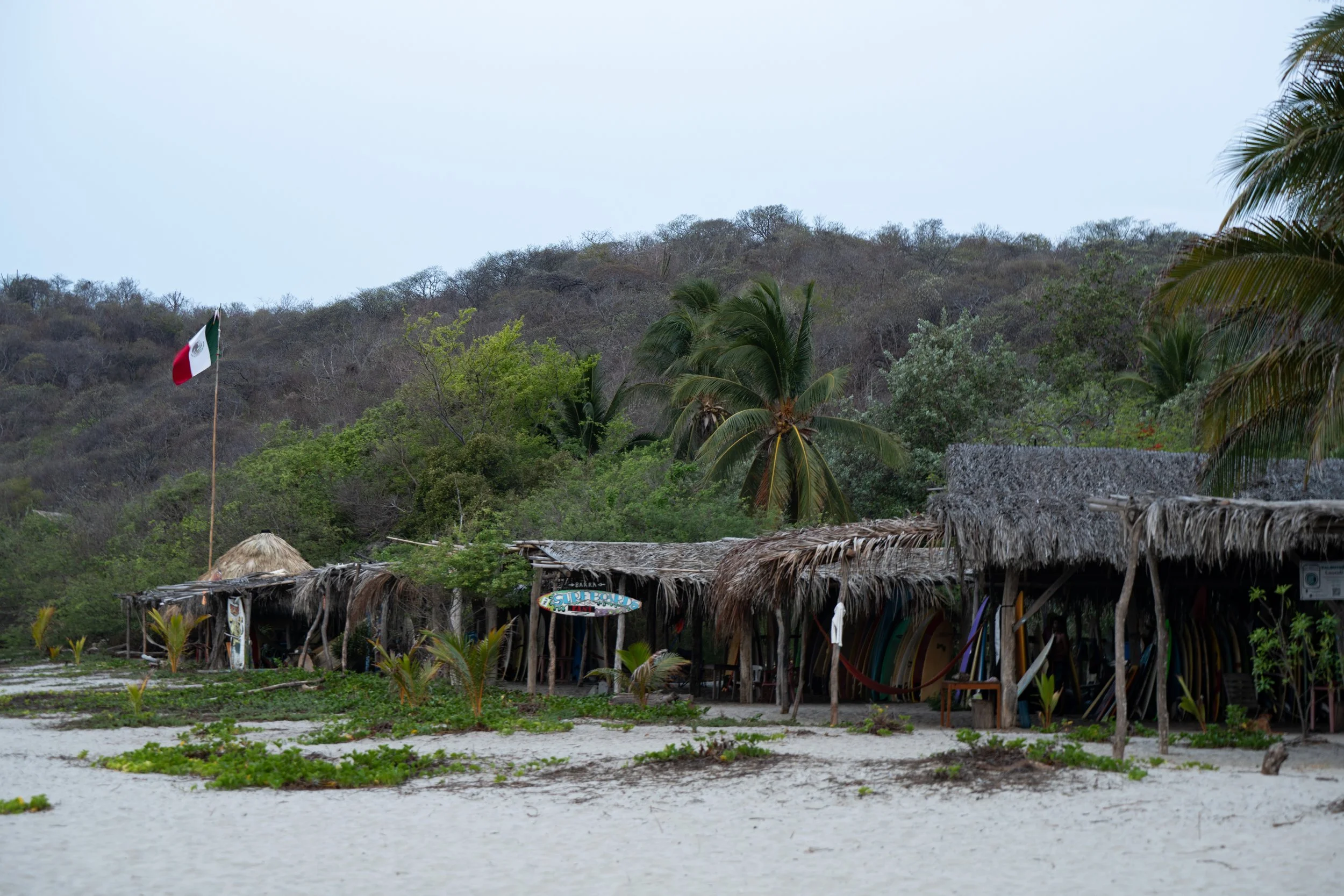 Beachside huts made of wood and thatch with a sign that reads 'Surf Bottle' in front, surrounded by lush greenery, palm trees, and hills in the background with an overcast sky.