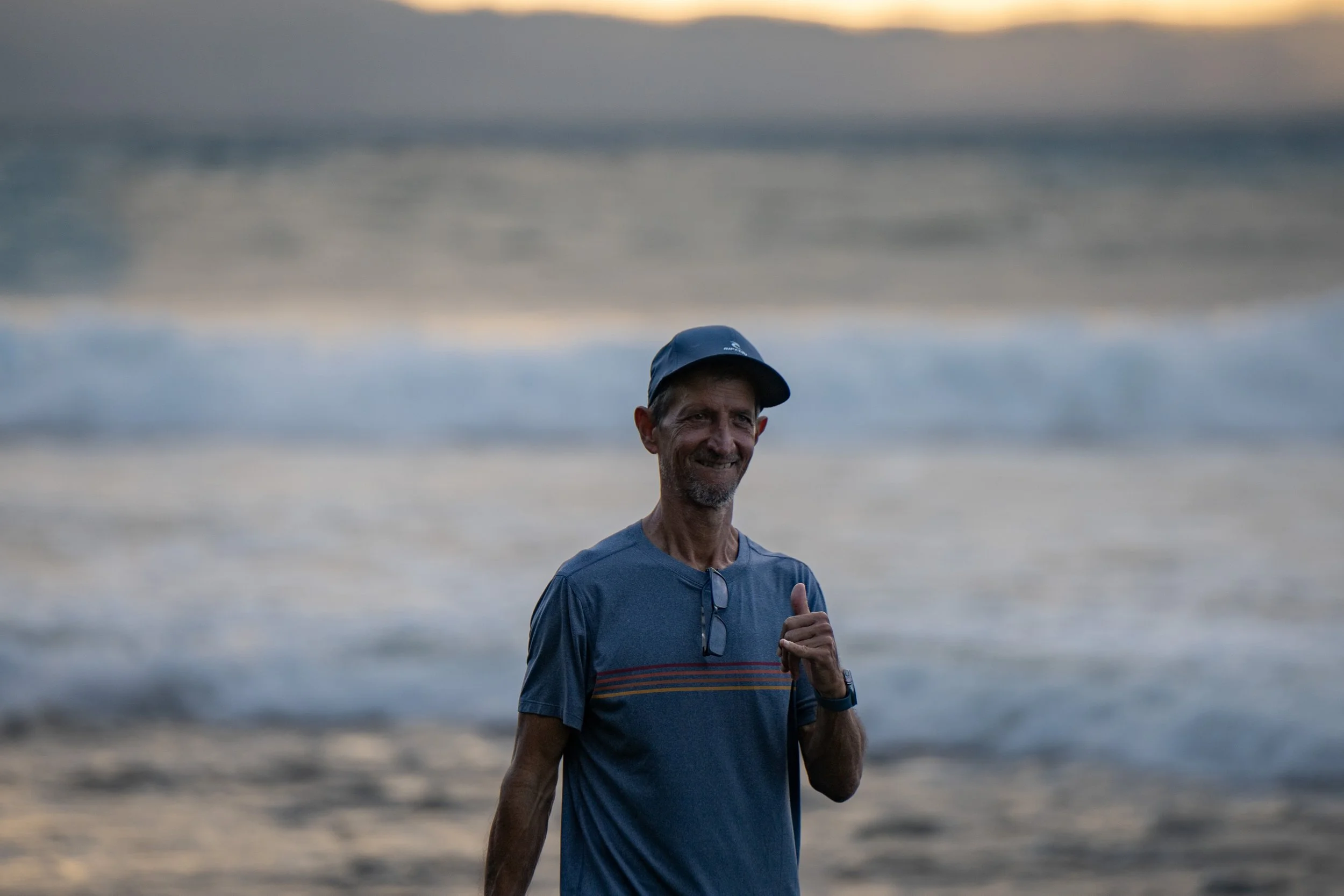 A man in a blue T-shirt and cap giving a thumbs up at the beach during sunset, with ocean waves and cloudy sky in the background.