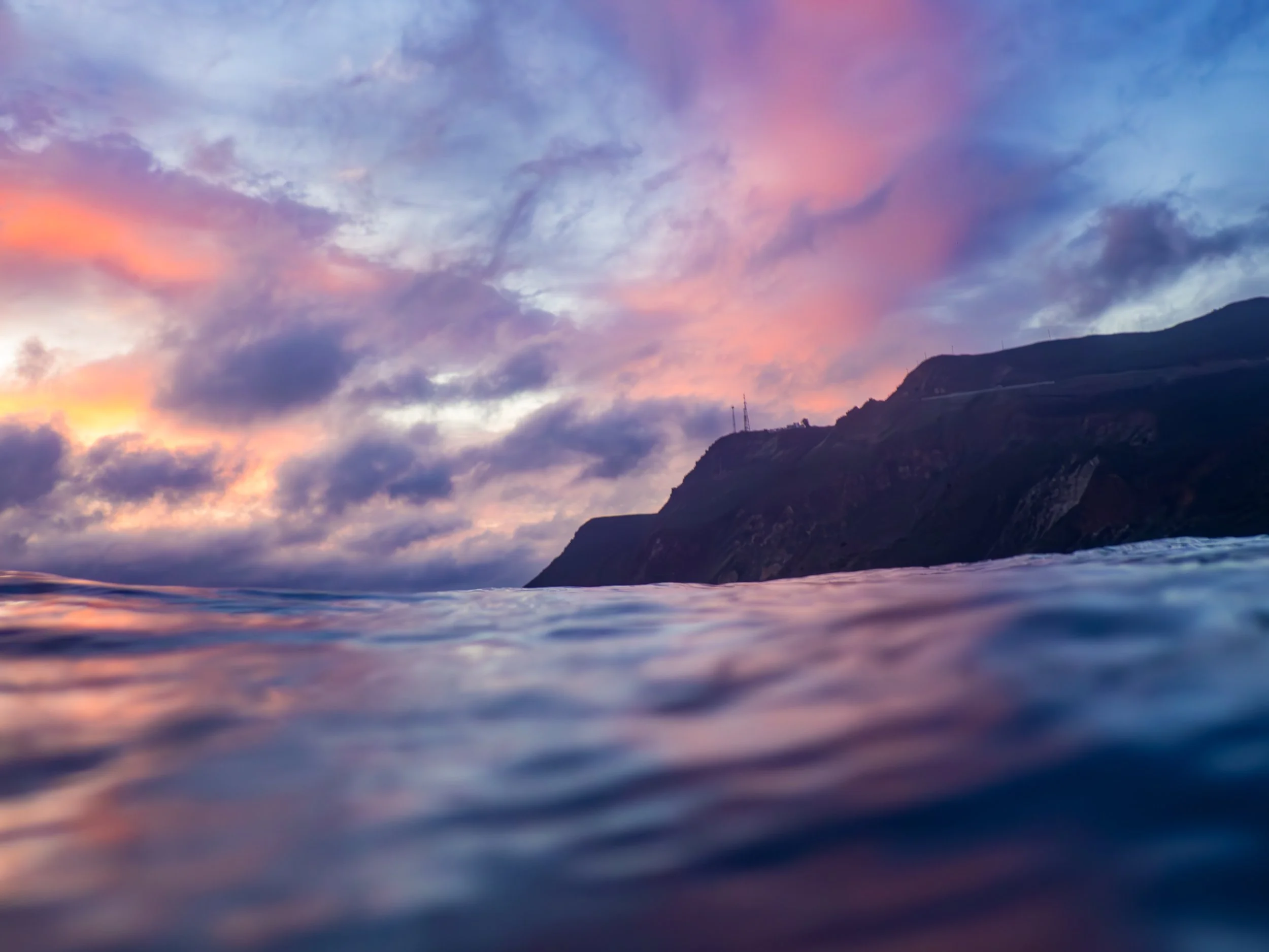 A view of the ocean with purple and pink clouds in the sky and a mountain in the distance.