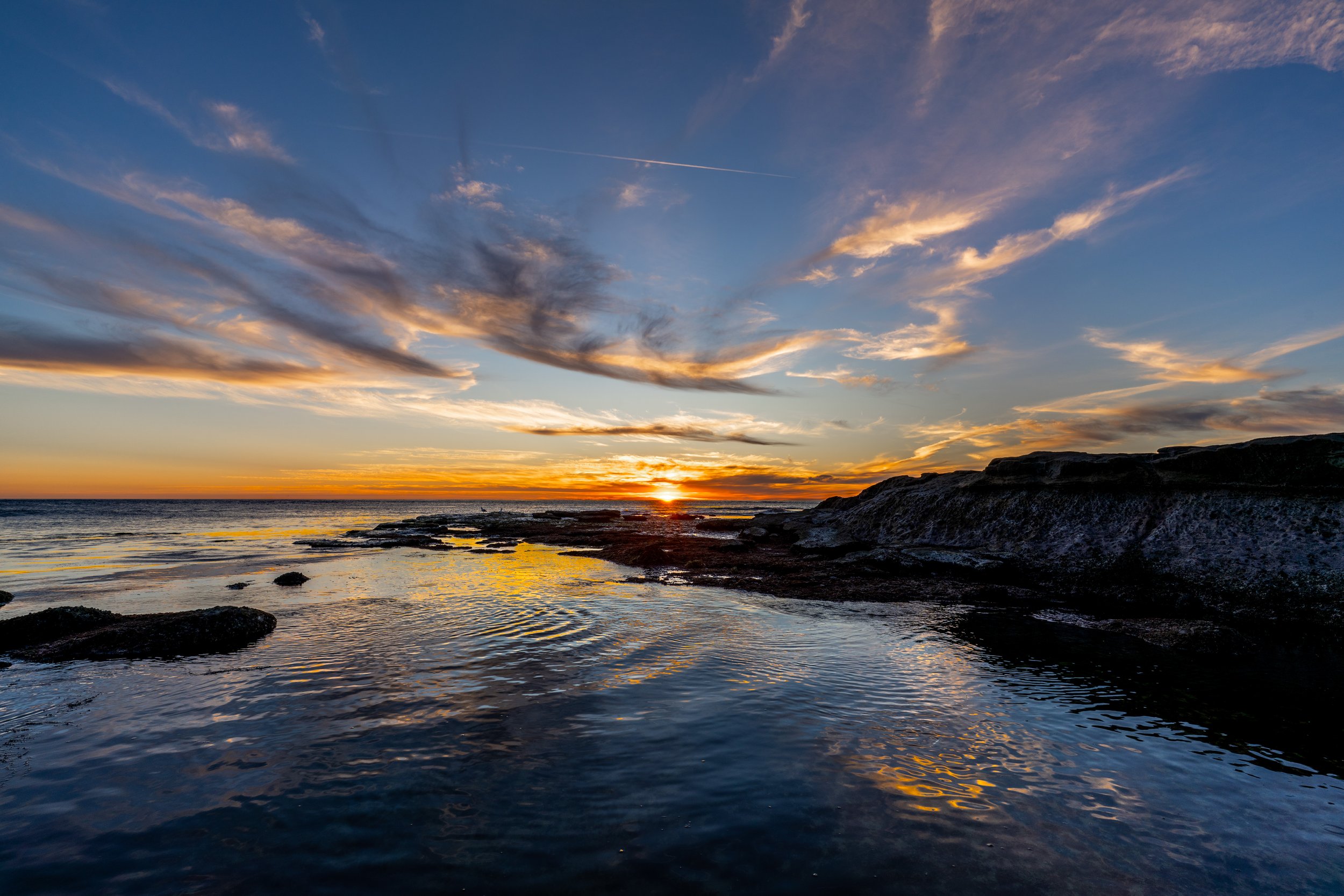 Sunset over the ocean with clouds in the sky and rocks in the foreground.