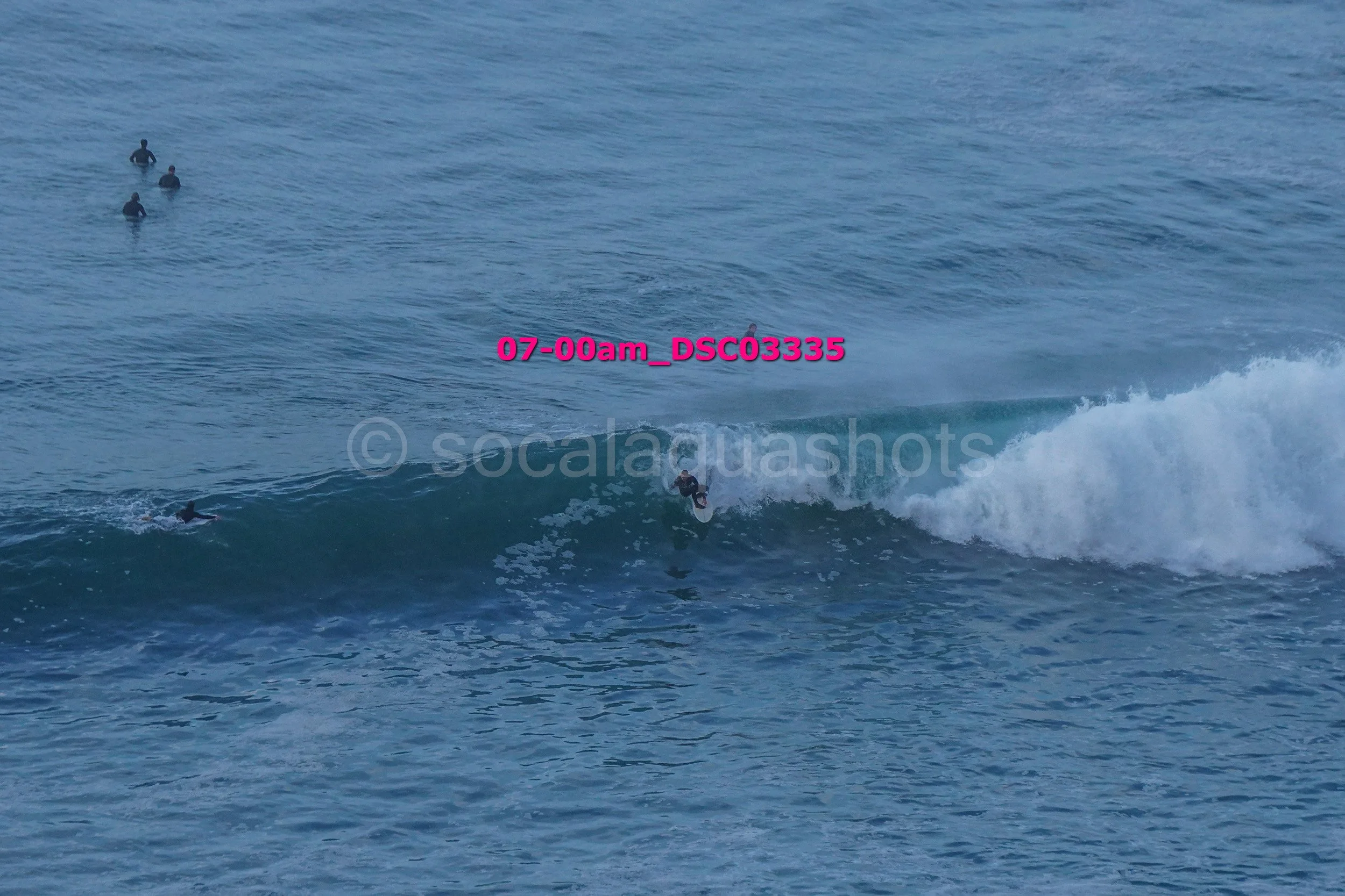 A person surfing on a wave with four other surfers in the water nearby, in the ocean with visible surf.