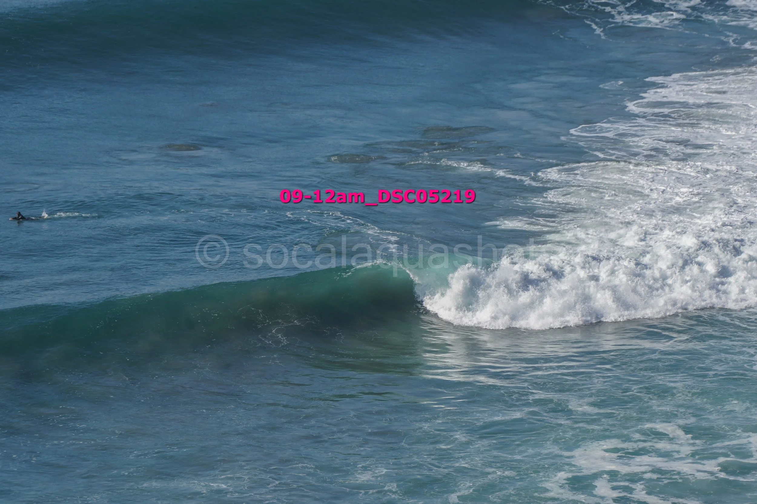 A surfer riding a turquoise wave in the ocean, with a swimmer paddling nearby.