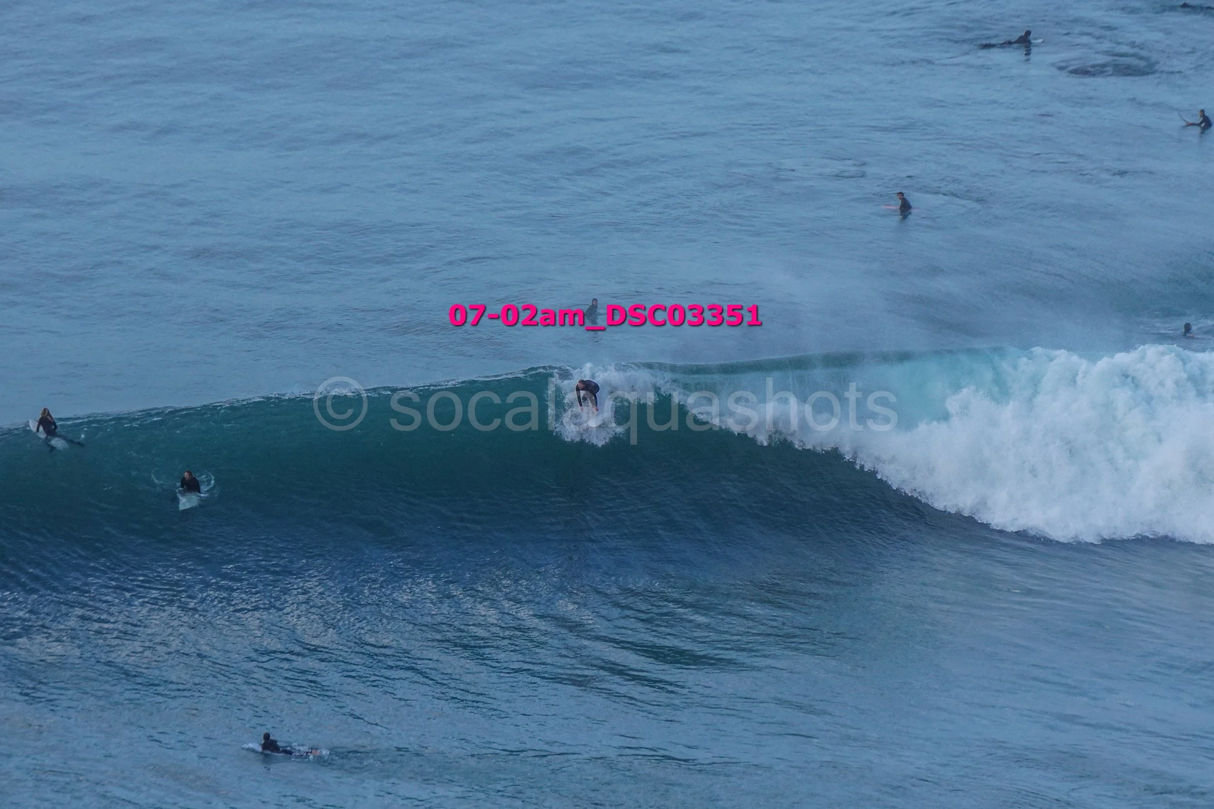 Surfer riding a wave with several other surfers in the water, ocean scene in daylight.