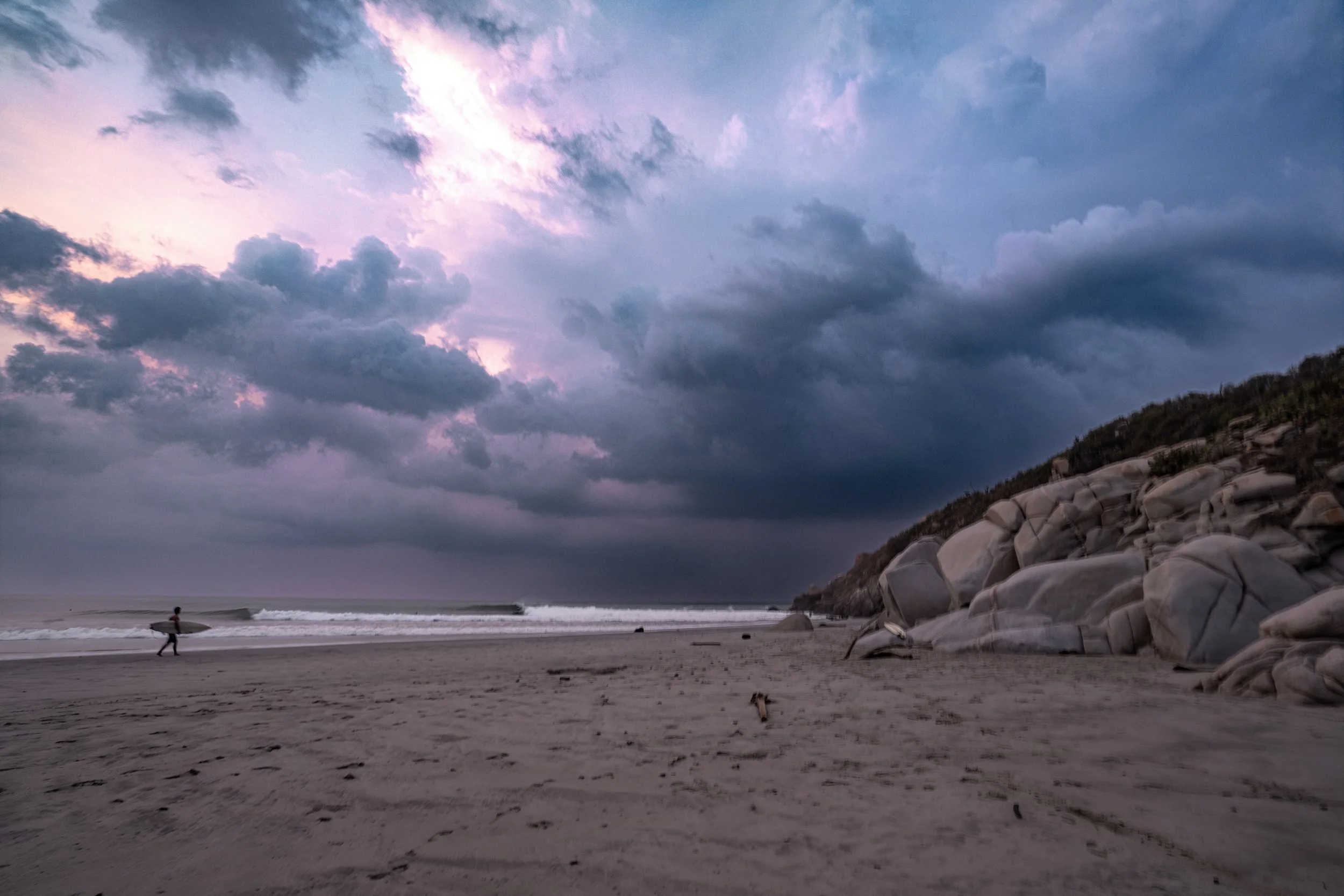 Person walking with a surfboard along the sandy beach during cloudy evening with a purple sky and rocky hillside in the background.