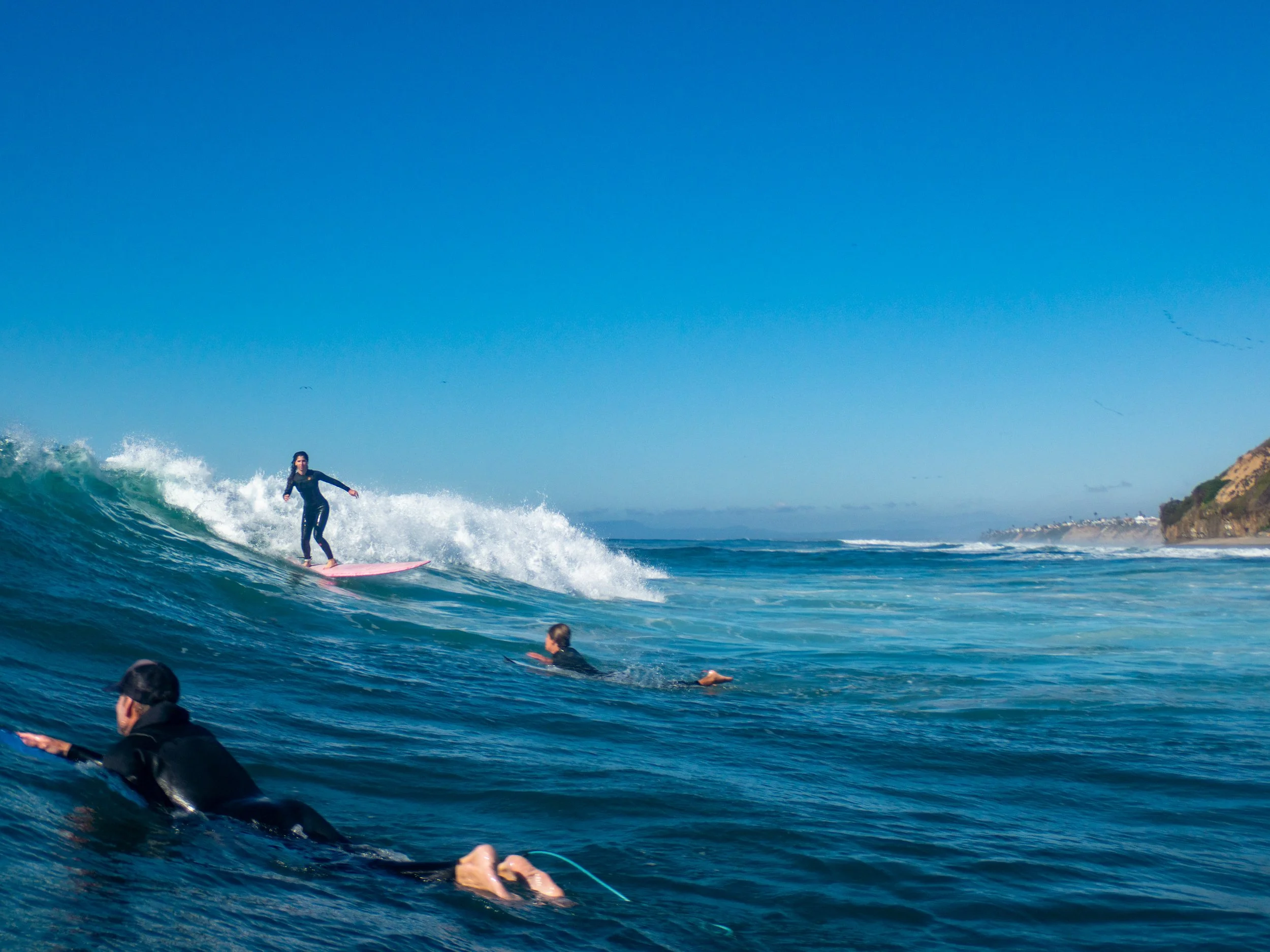Surfer riding a wave while several people swim in the water nearby on a sunny day at the beach.