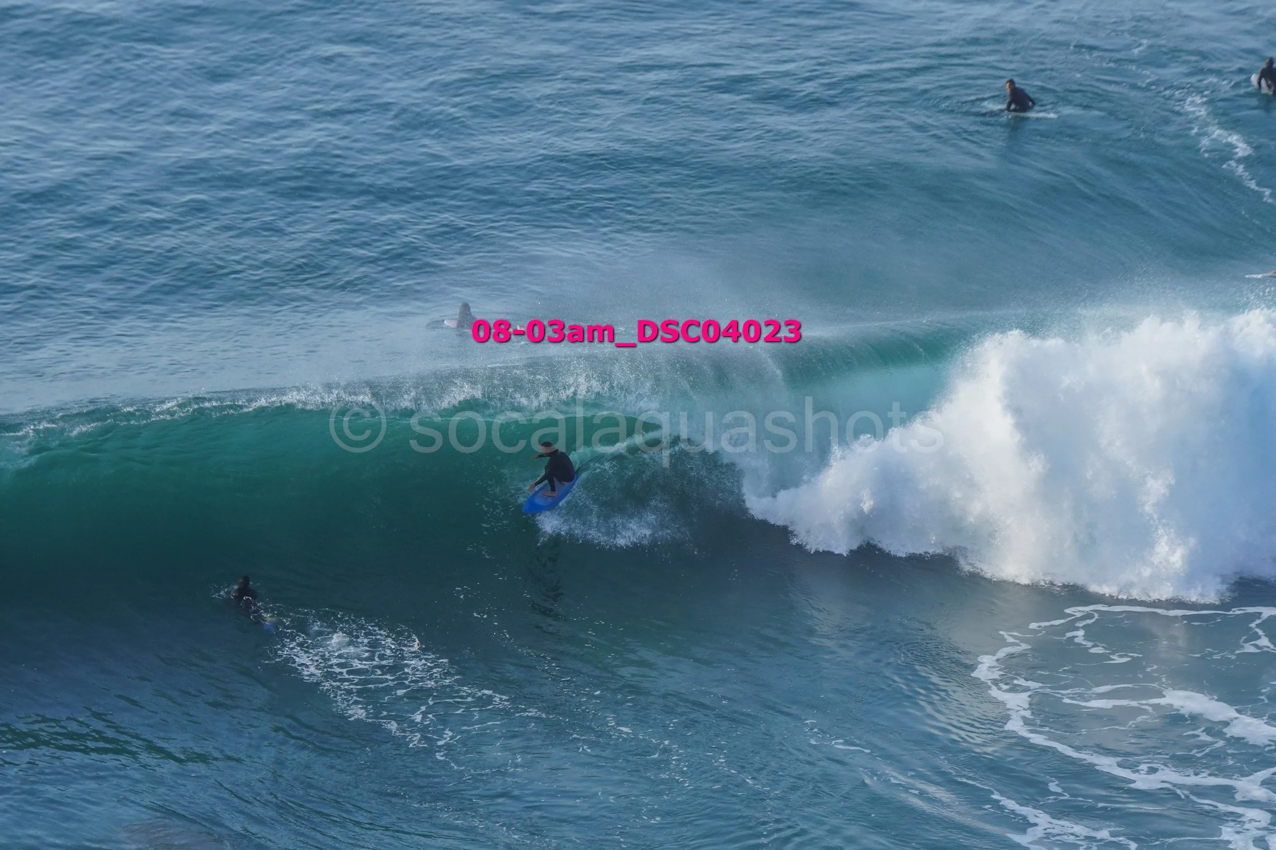 Surfer riding a large wave with several people in the water and others on surfboards in the background.