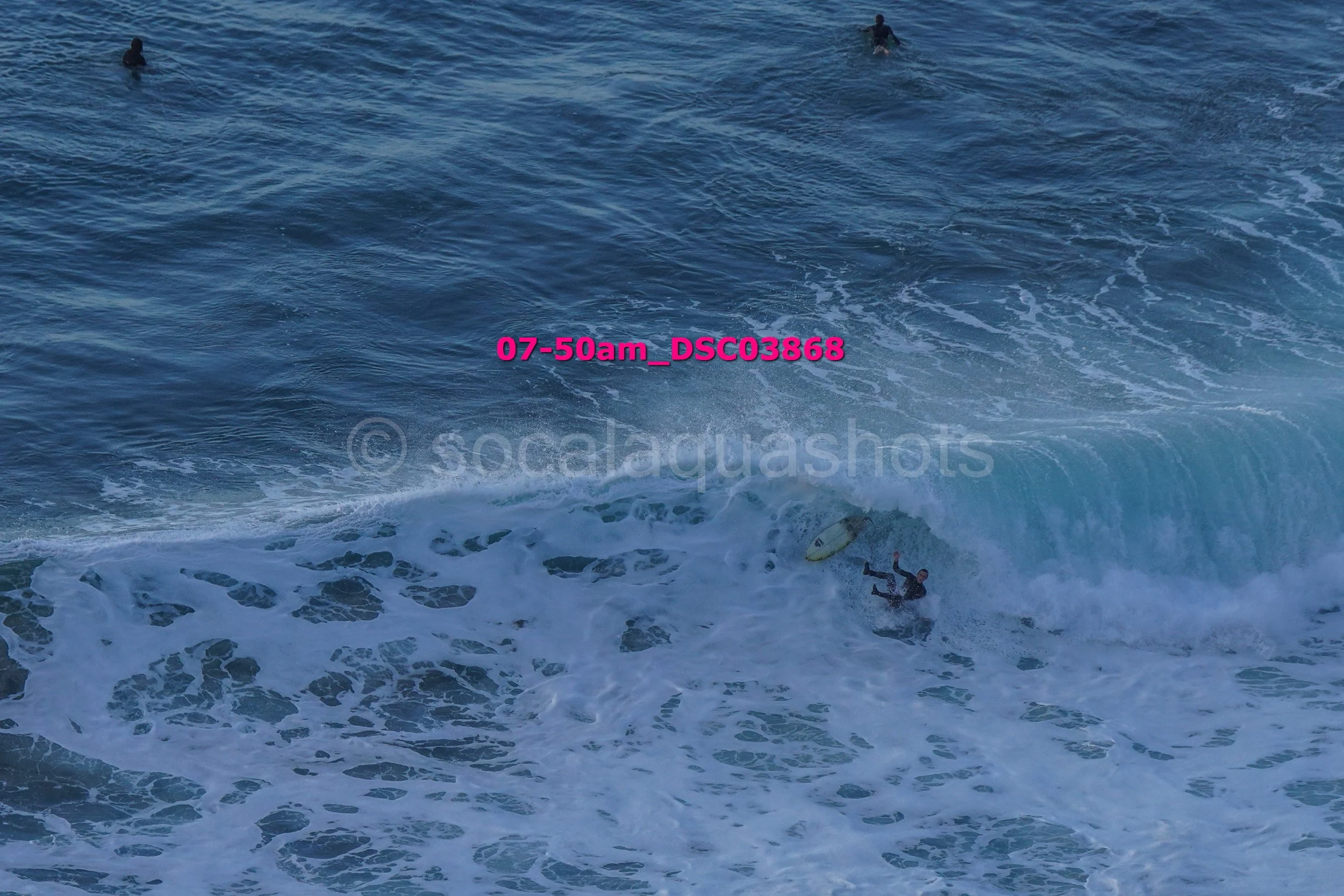 A surfer falling off his surfboard in the ocean waves with other surfers in the background.