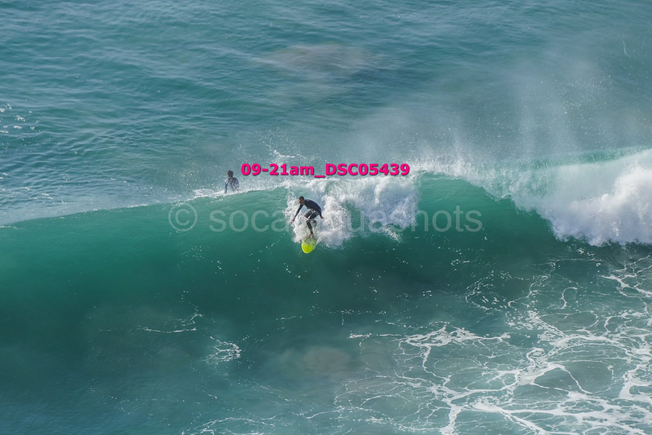 A person surfing on a large green wave with another surfer visible in the background in the water.