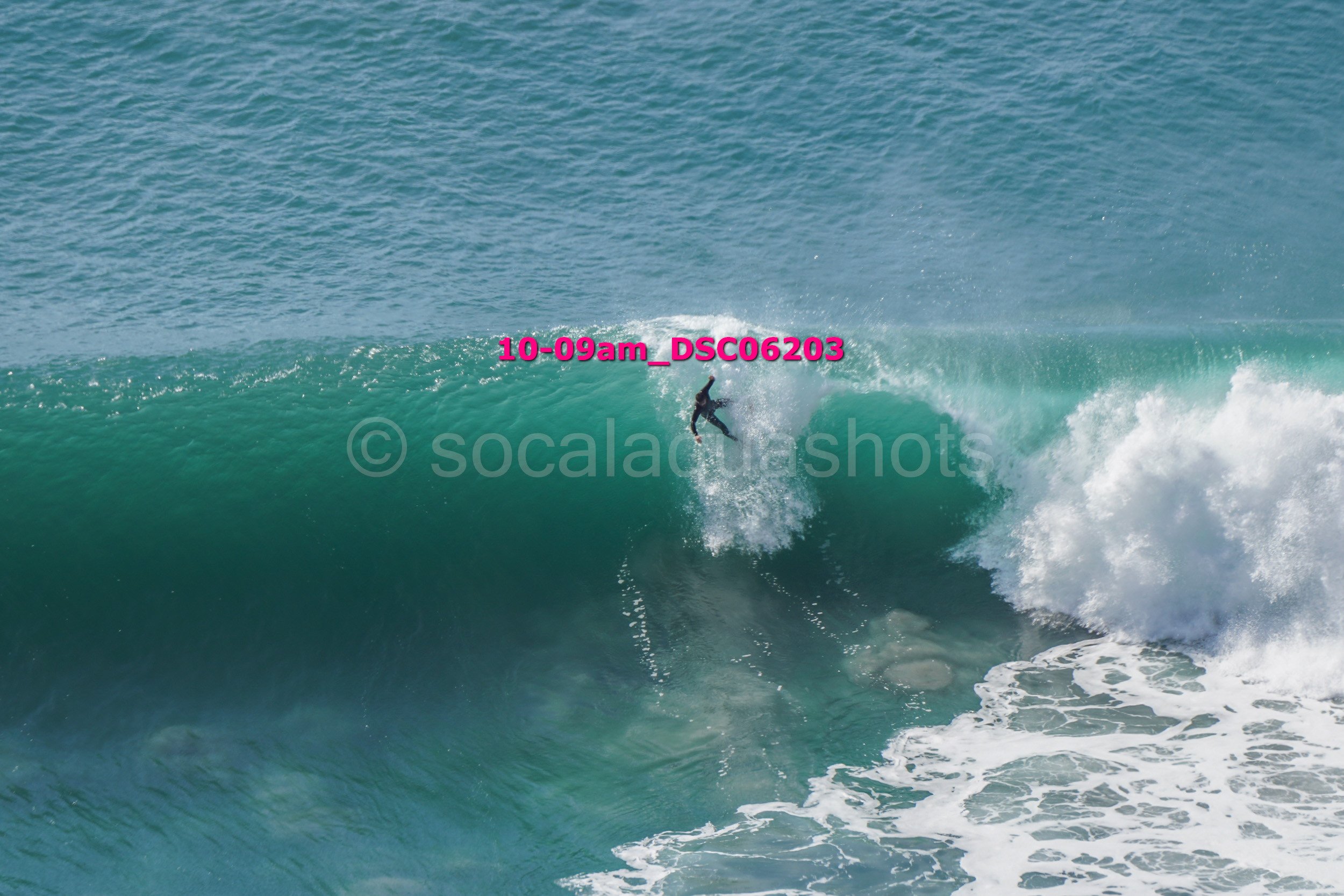 A surfer riding inside a large ocean wave with a visible white surf foam, turquoise water, and an open sky in the background.