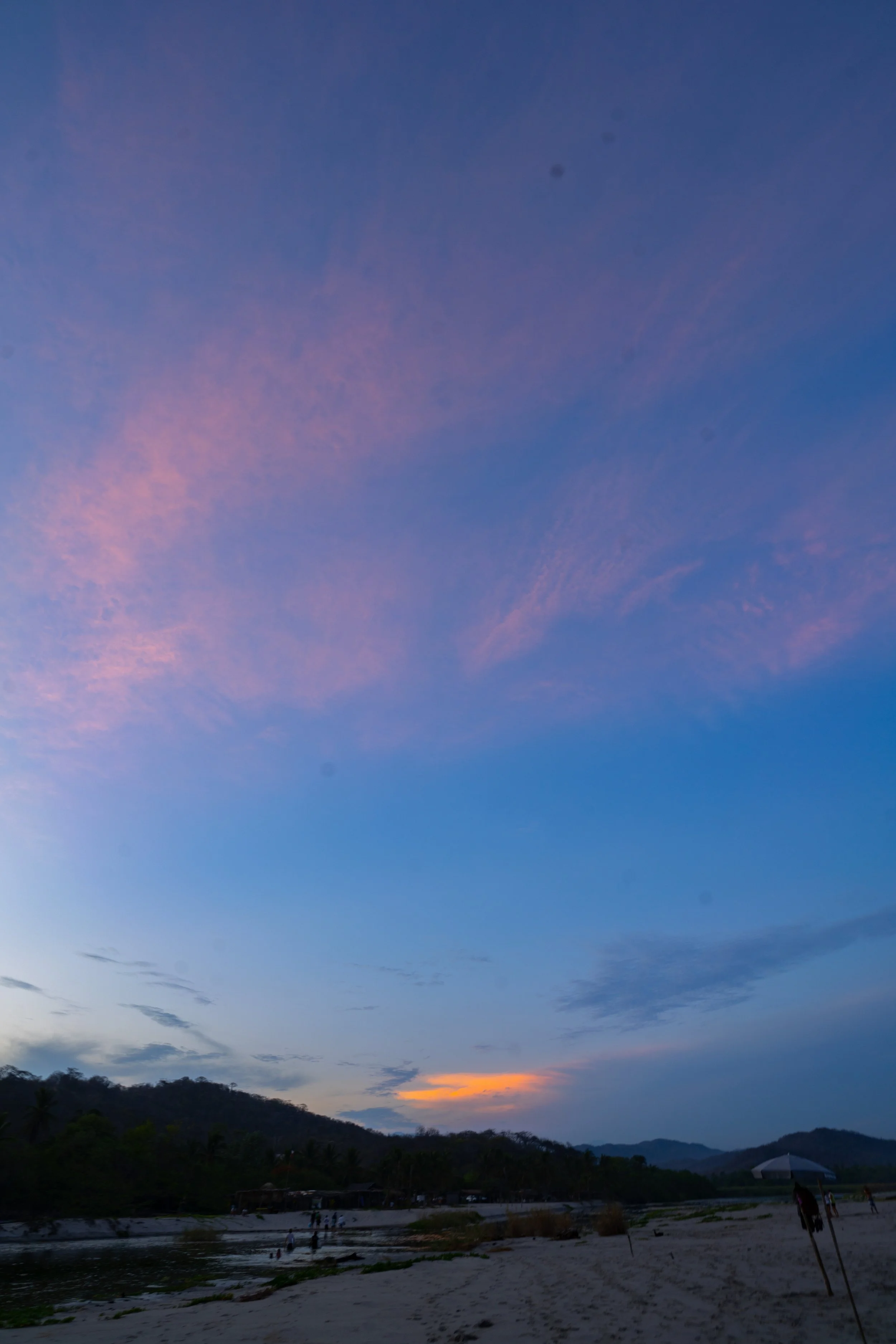 A beach at sunset with a sky filled with blue and pink clouds, silhouetted hills in the distance, and people near the water and on the sand.