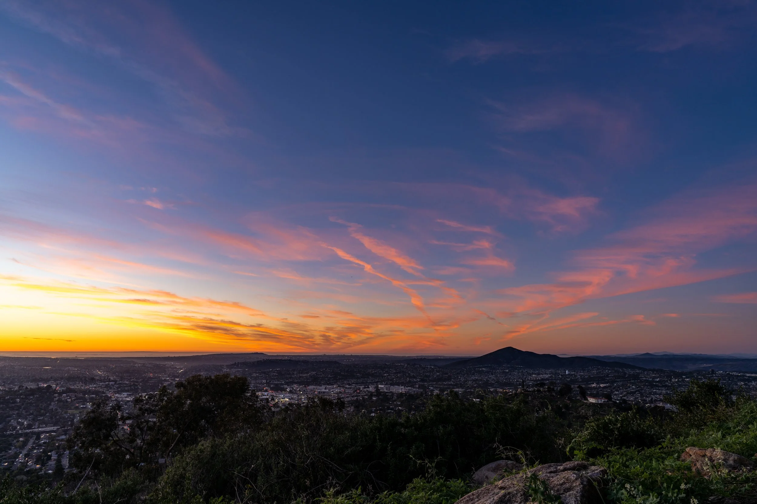 Sunset over a city with colorful clouds in the sky and a mountain in the distance. Foreground shows trees and rocks.