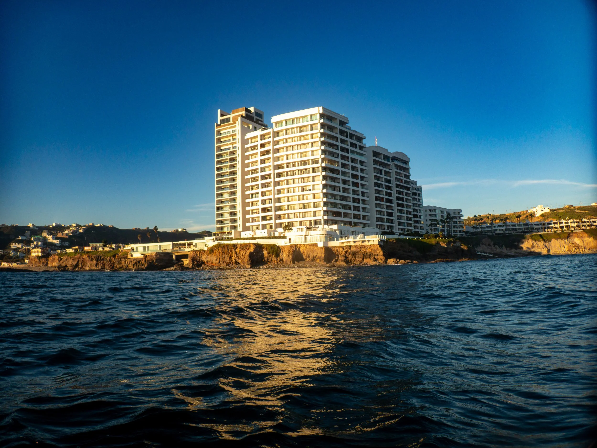 Modern high-rise building on rocky shoreline near water with a clear blue sky in the background.
