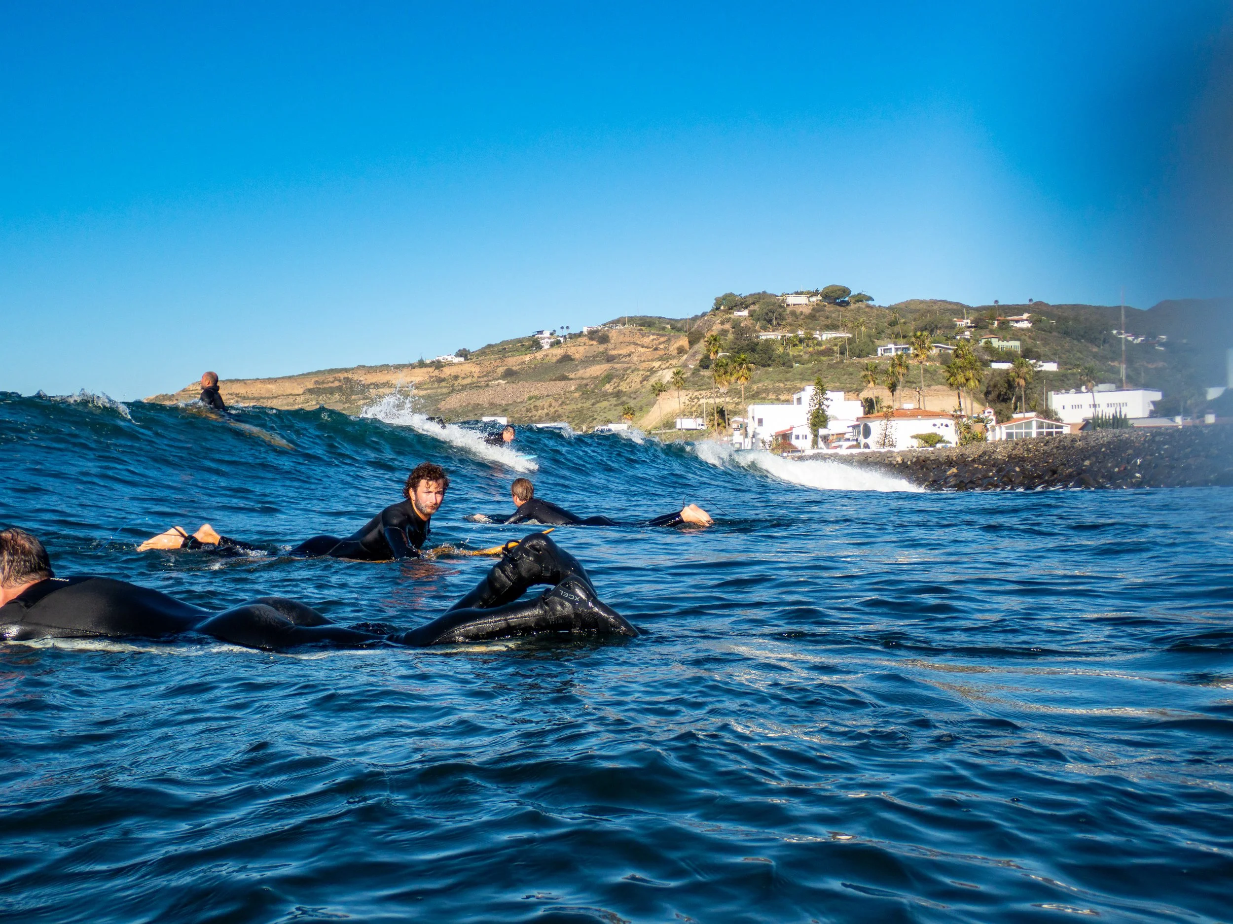 People in wetsuits swimming in the ocean with a coastal hillside and white houses in the background.