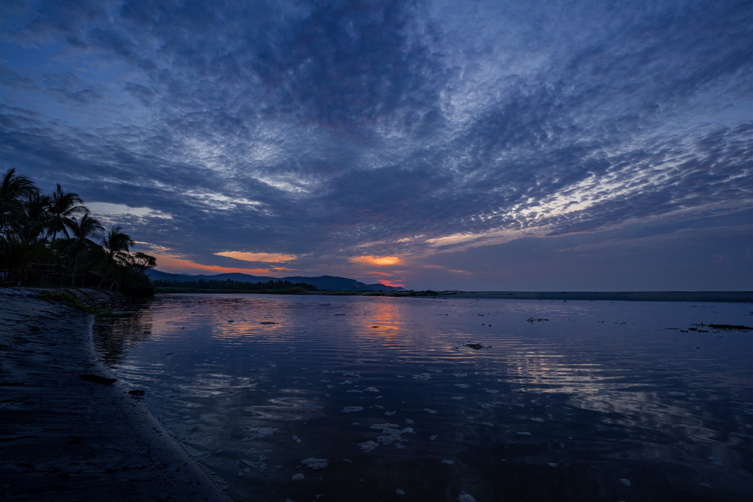 A sunset over a river with a cloudy sky and silhouettes of palm trees on the left bank.