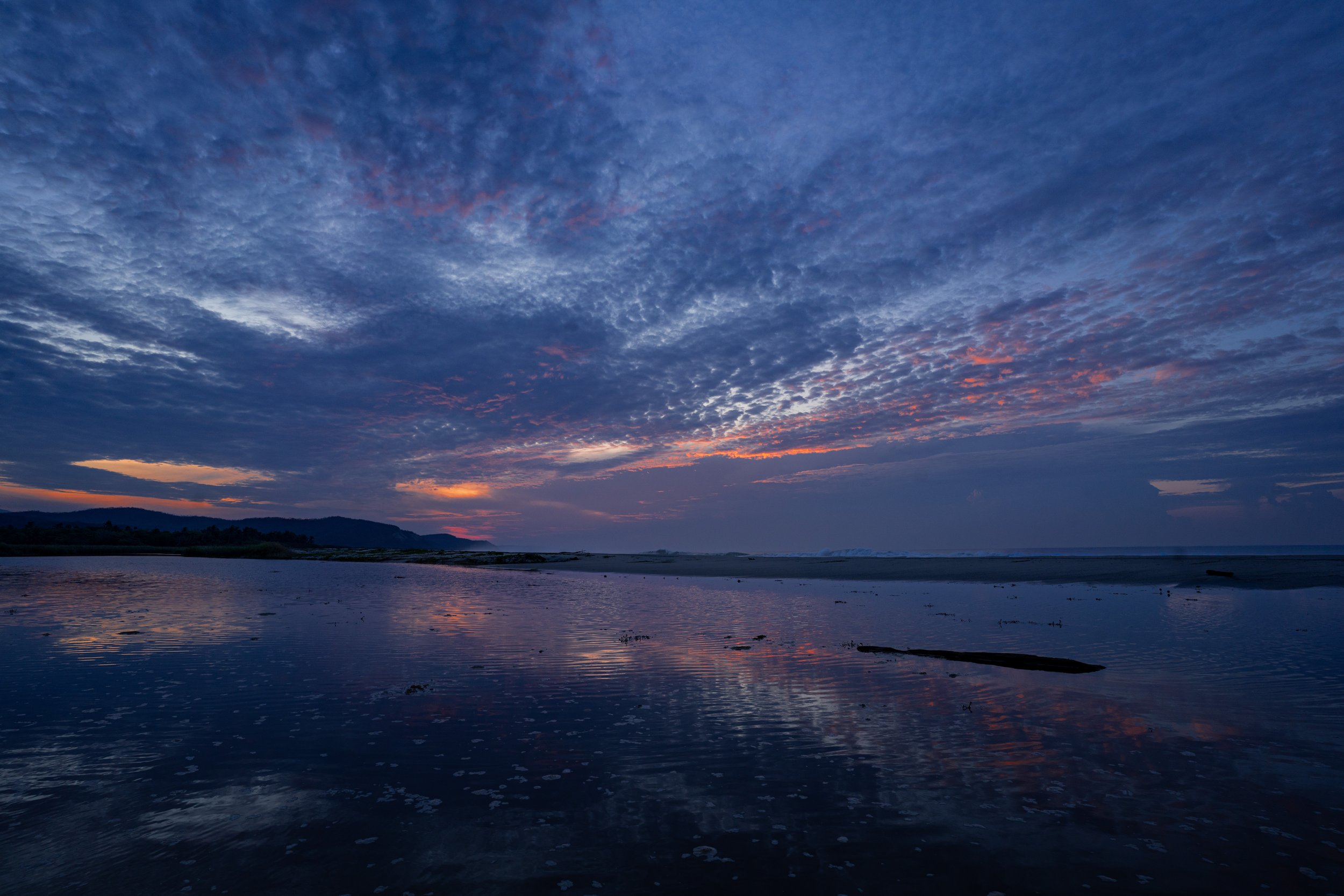 Ocean or large water body at sunset with clouds and a mountainous coastline in the distance.