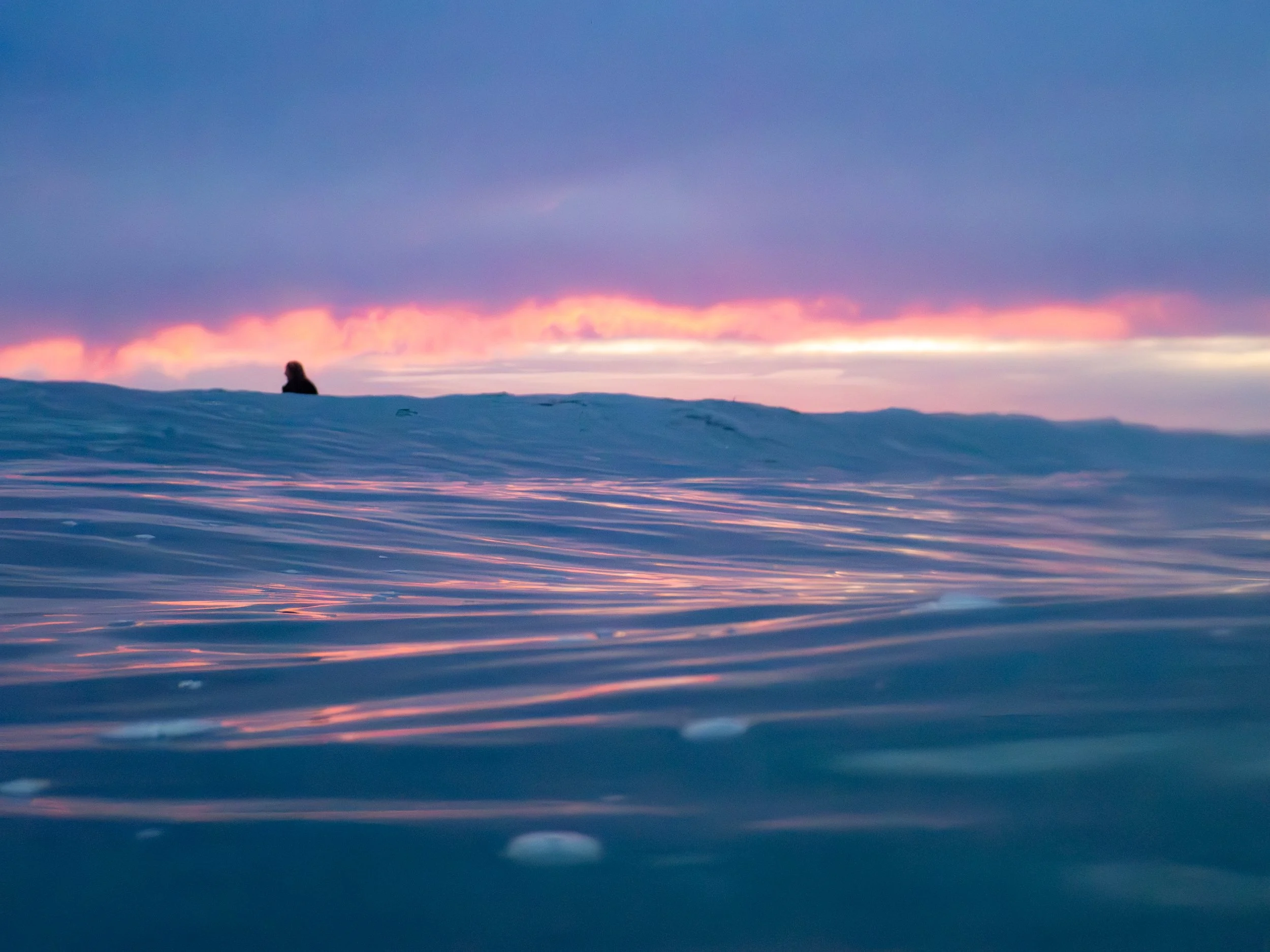 A person sitting on a snow-covered landscape at sunset with a colorful sky and calm water in the foreground.