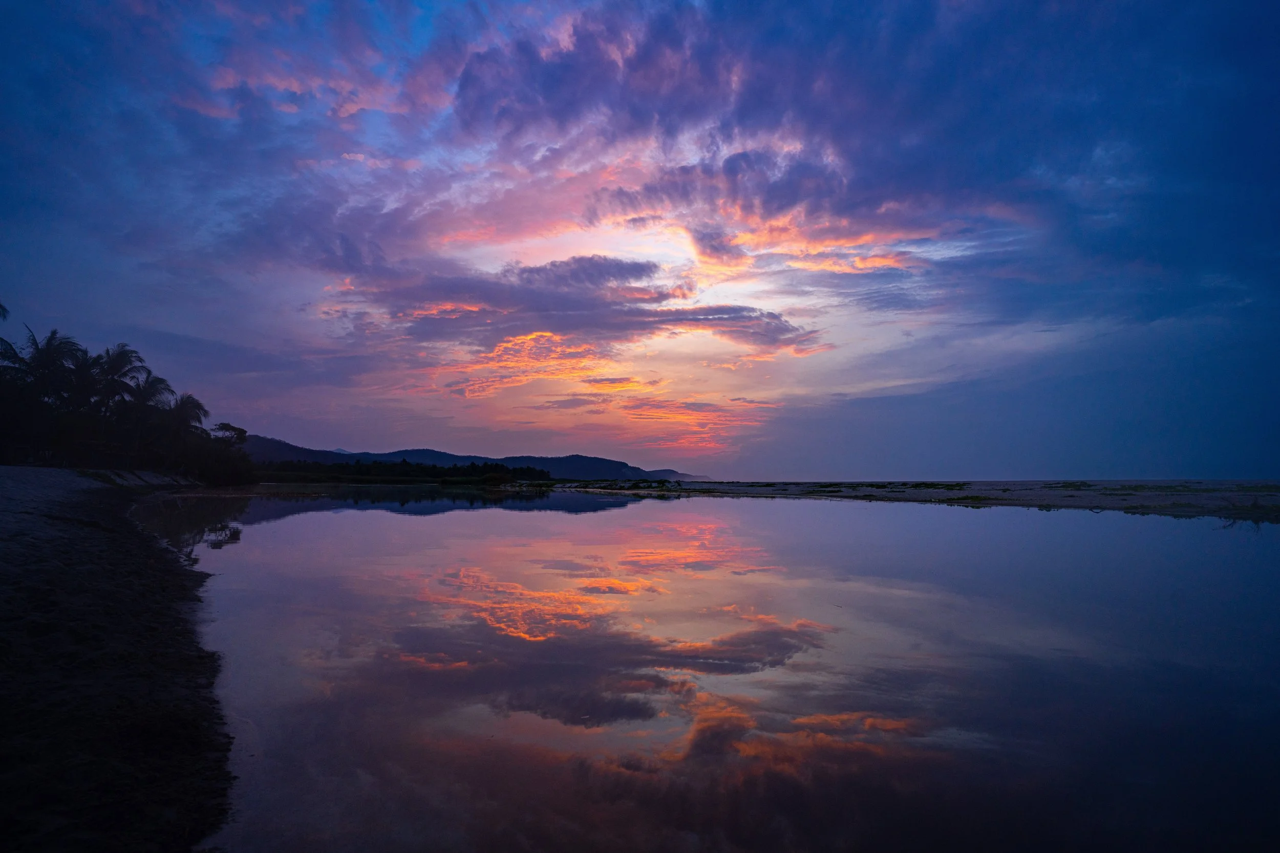 Sunset over a calm river with colorful sky and clouds, silhouettes of trees and hills in the distance.