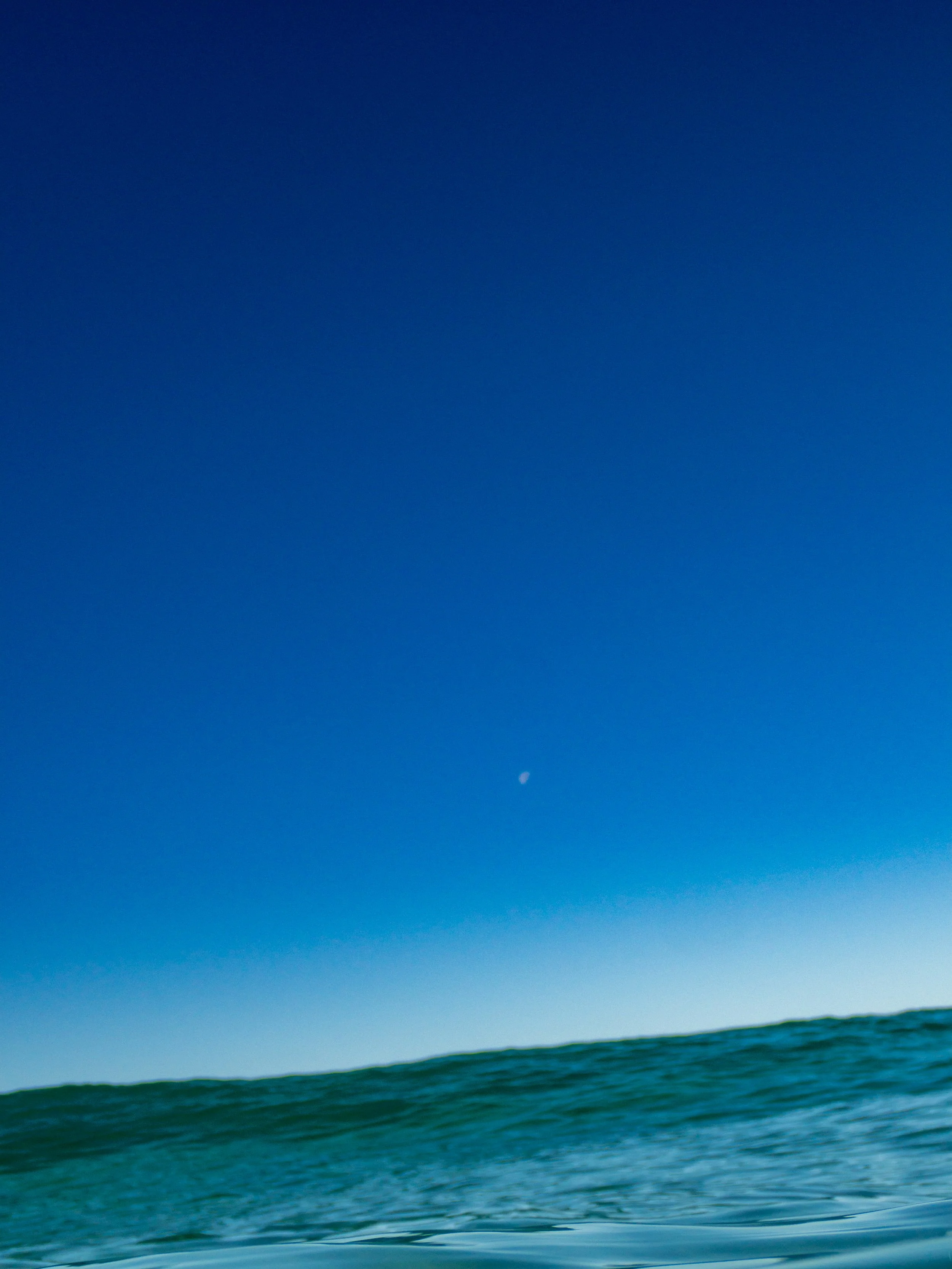 View of ocean waves under a clear blue sky with a small visible moon.