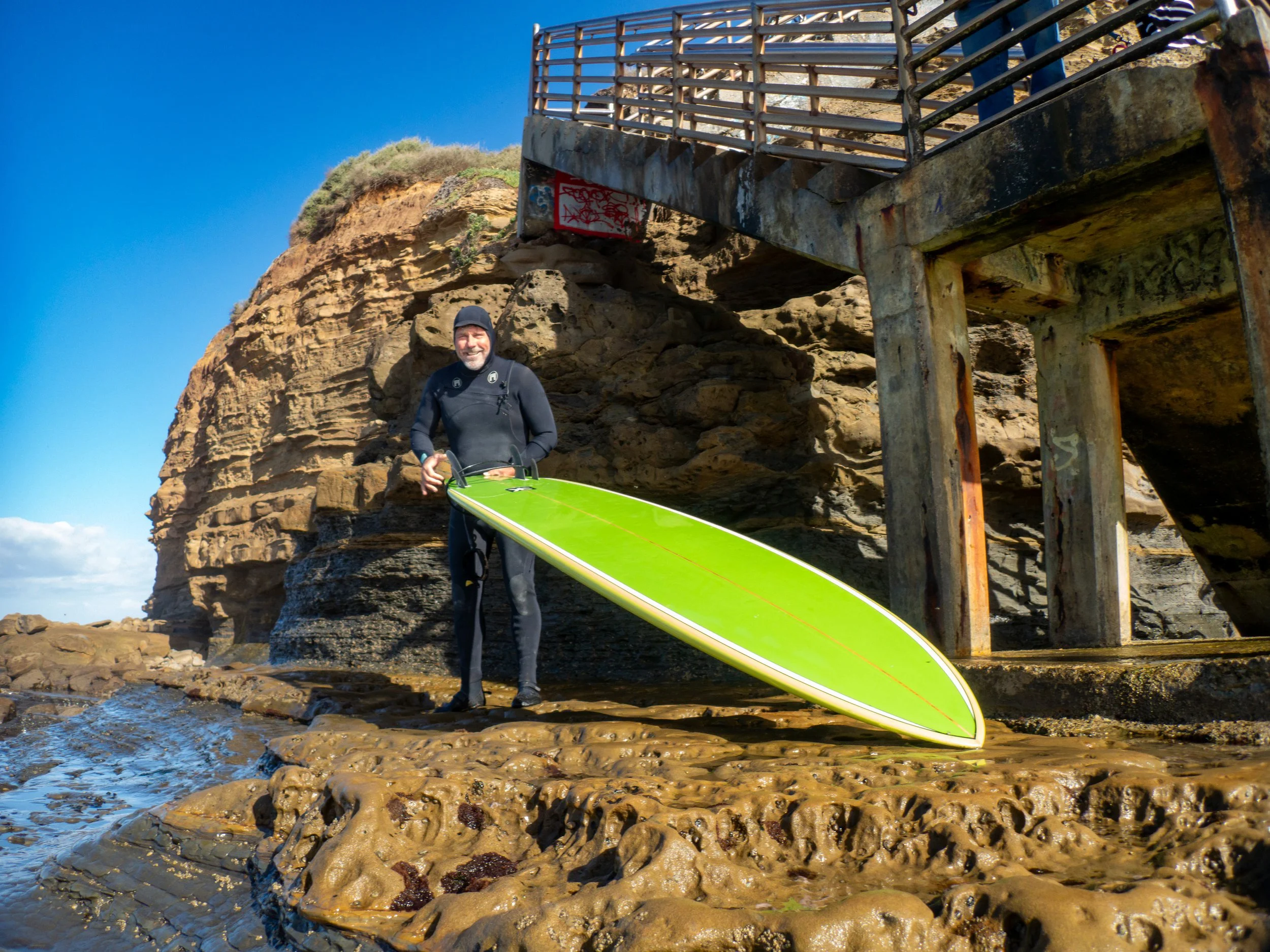A man in wetsuit standing next to a surfboard on rocky beach under a large cliff and a staircase structure.
