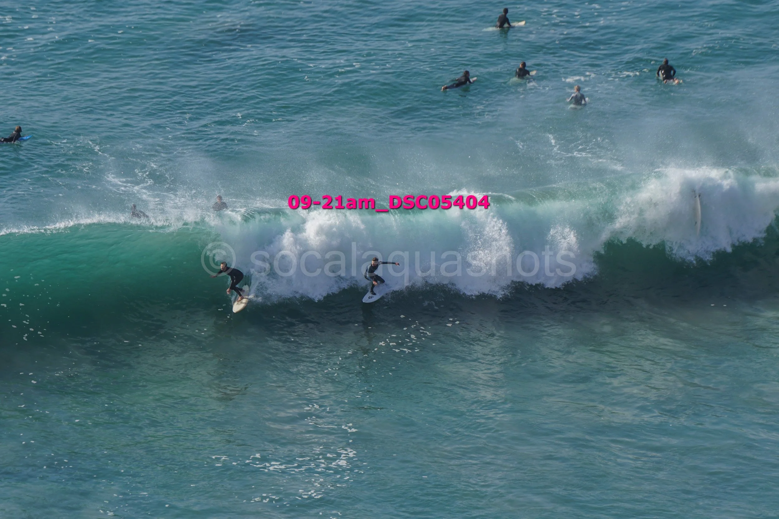 Several surfers riding and waiting for waves in the ocean, some on surfboards on a large wave, others swimming in the water, with a person paddling on a surfboard farther out.