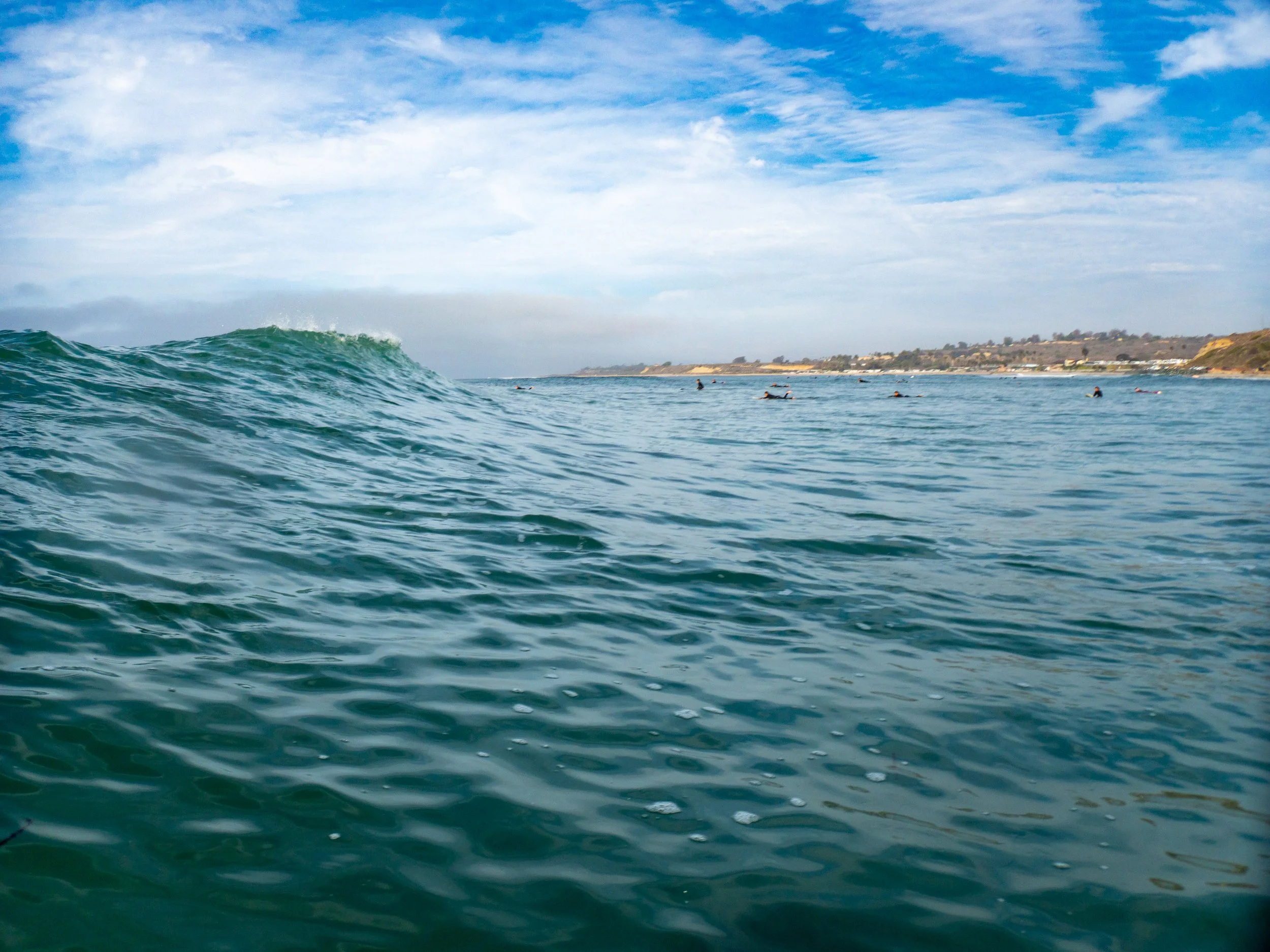 Ocean waves with surfers in the distance under a cloudy sky along a coastline.