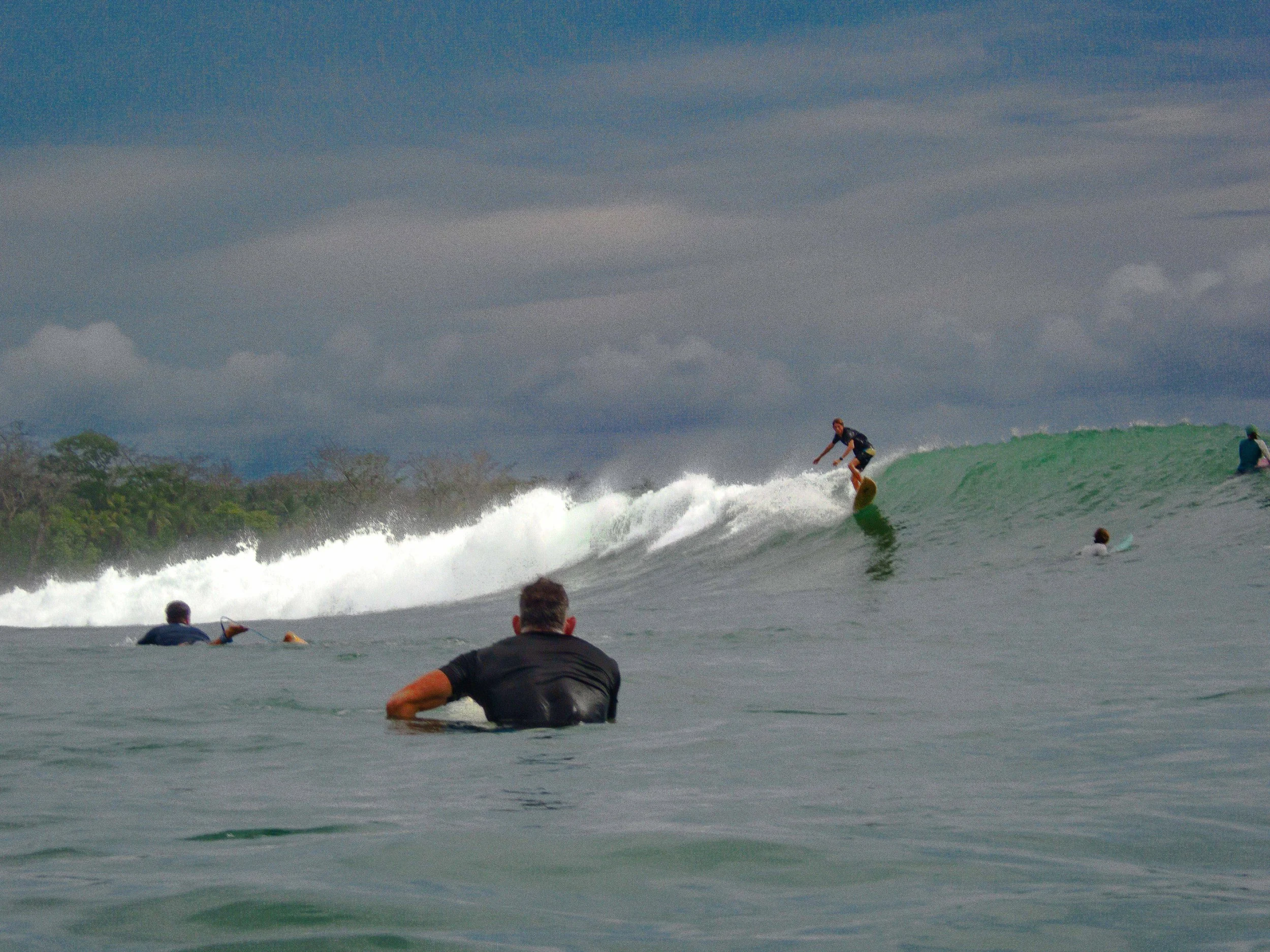 Surfer riding a wave with other surfers in the water under a cloudy sky.
