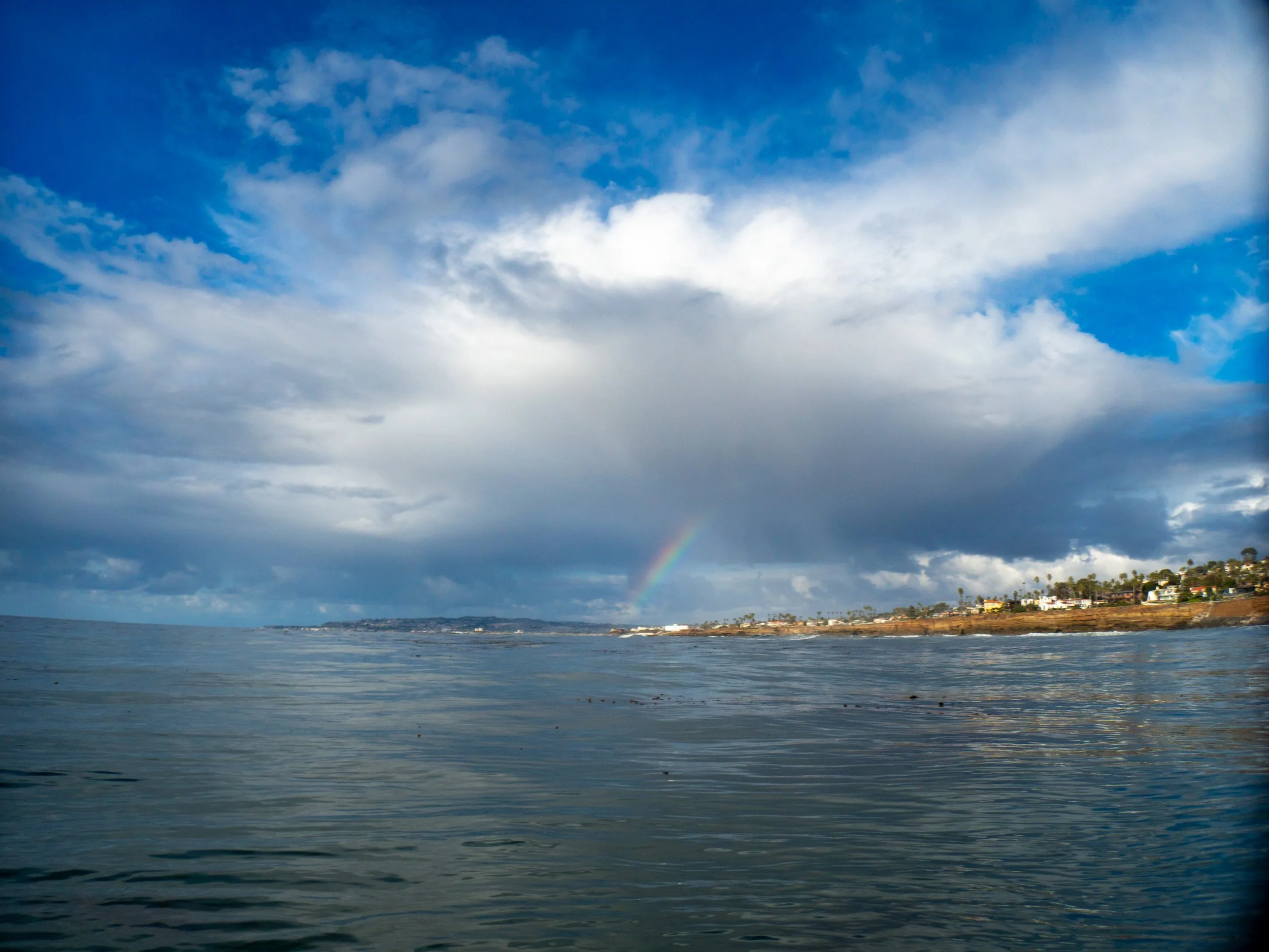 Seaside view with an ocean, cloudy sky with patches of blue, and a faint rainbow near the horizon. Coastal town with houses and trees on the right.