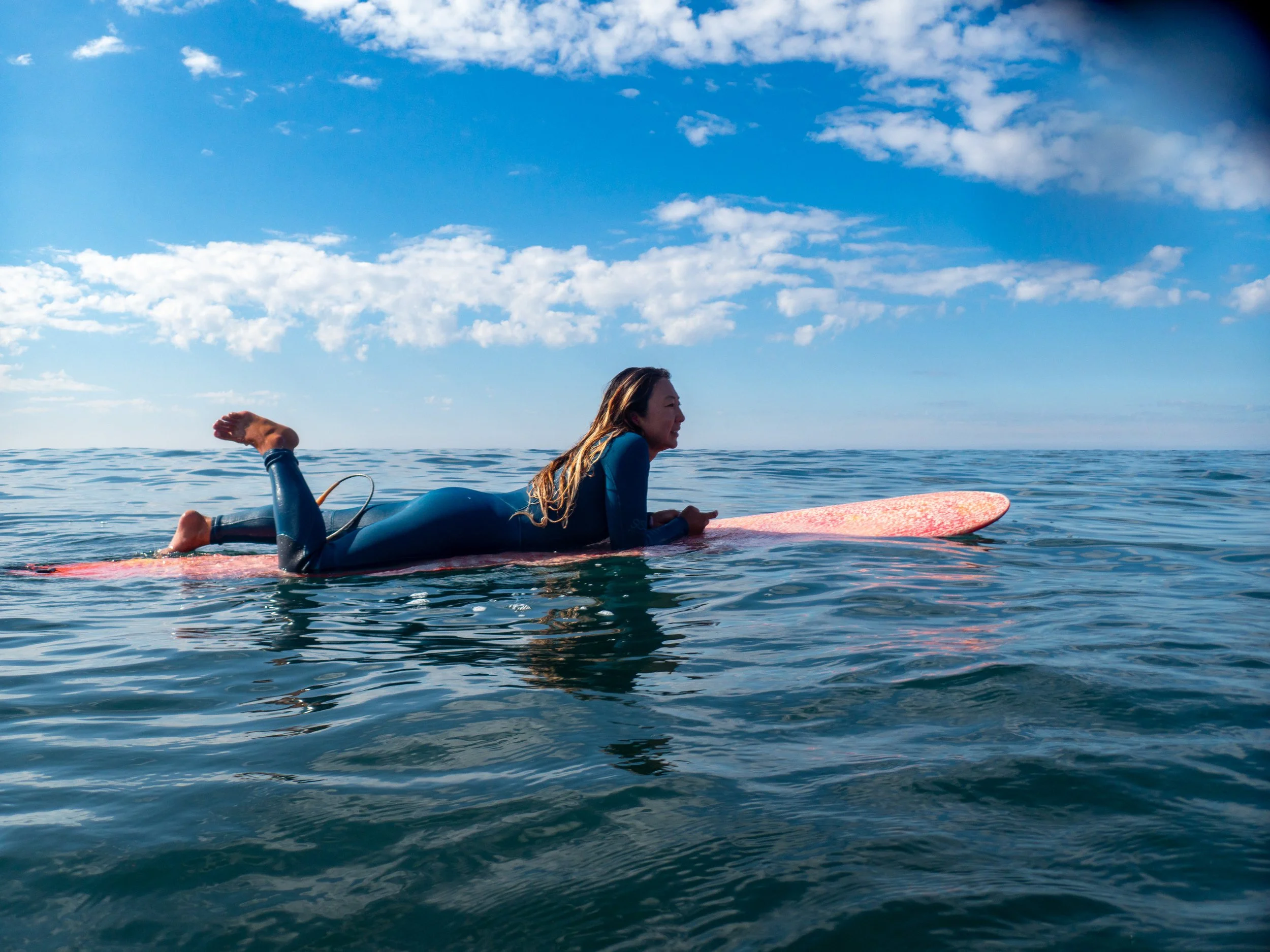 A woman in a wetsuit is lying on a pink surfboard on the water, looking forward with a blue sky and scattered clouds in the background.