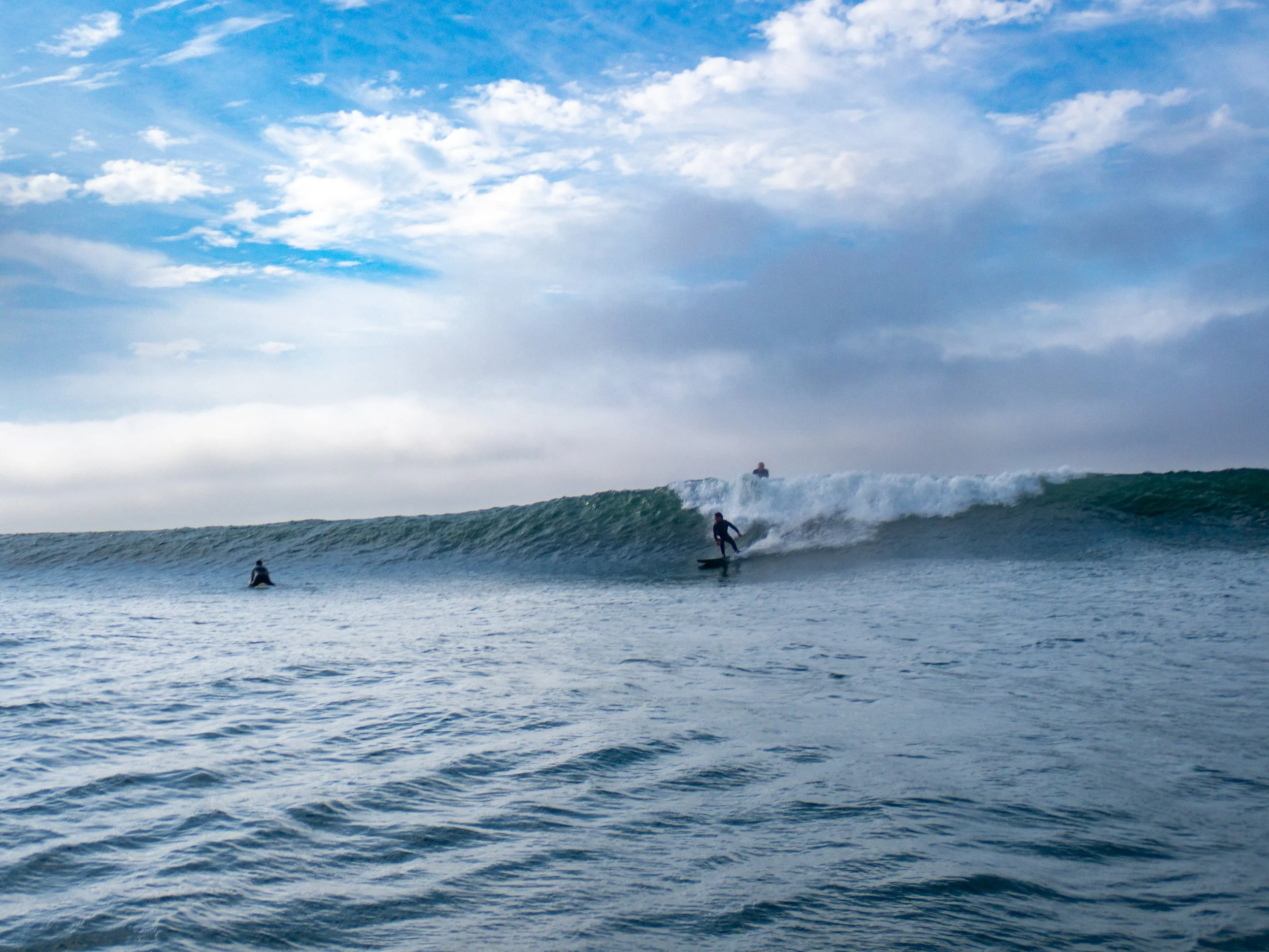 Surfer riding a wave in the ocean with two other surfers in the water and a partly cloudy sky overhead.