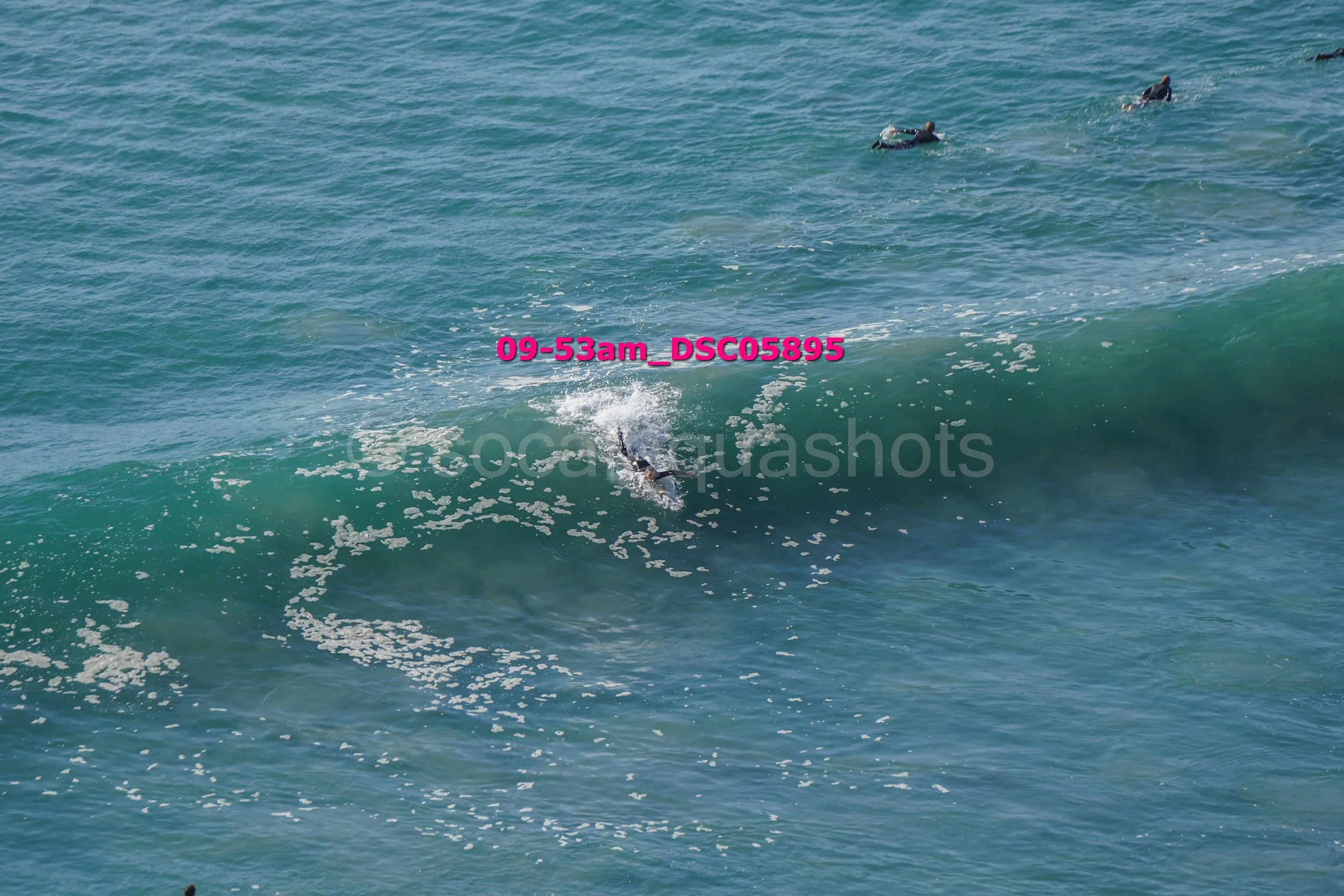 Surfer falling off surfboard into the ocean near a breaking wave with a few other surfers further out at sea.