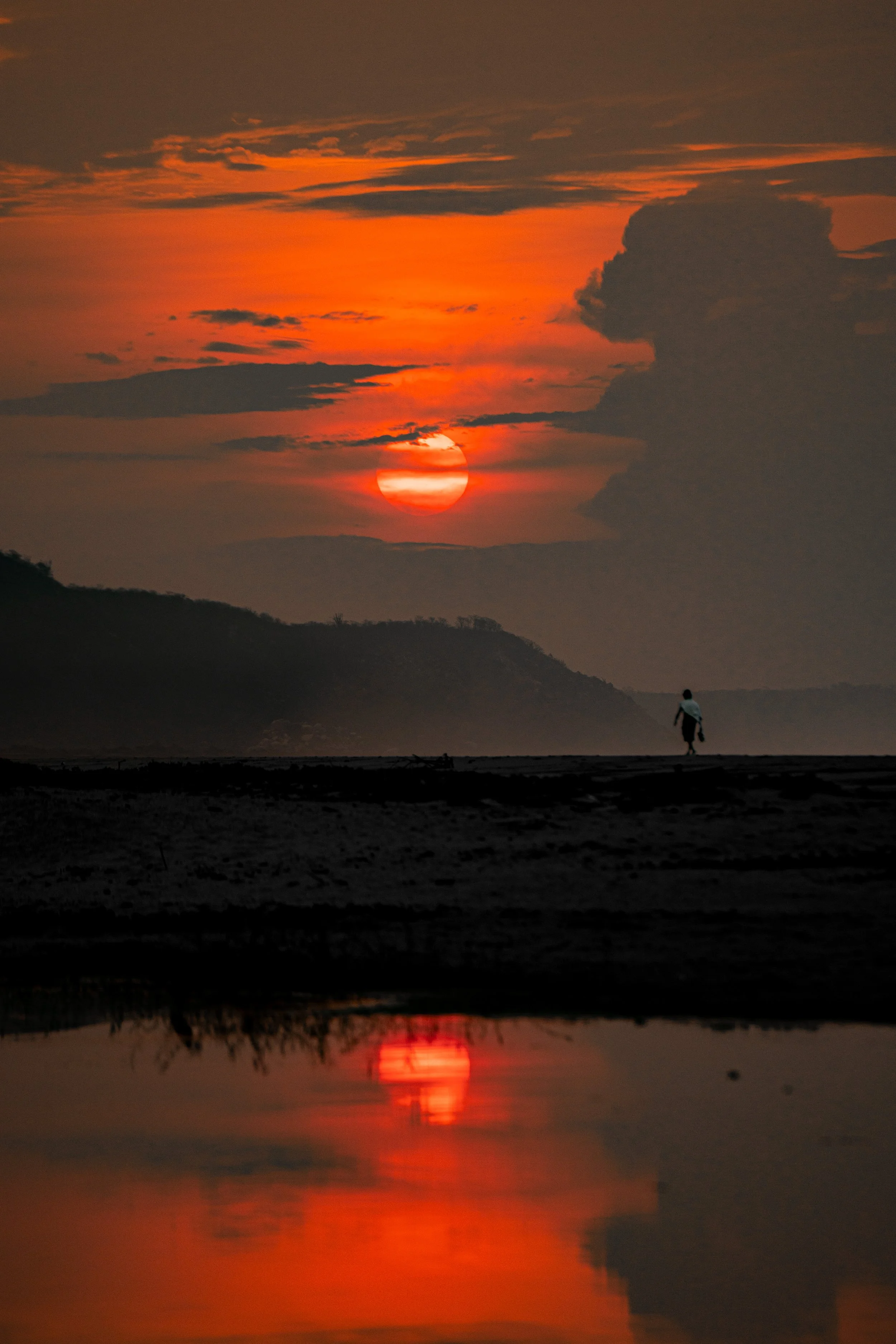 Sunset over mountains with orange and red clouds, a person's silhouette walking along a riverbank, and a reflection of the sunset on the water.