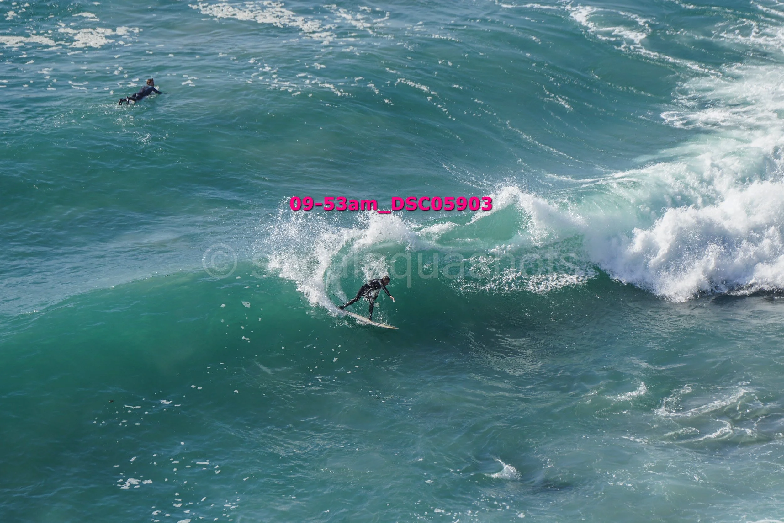 A surfer riding a large wave in the ocean, with another person floating in the water in the background.